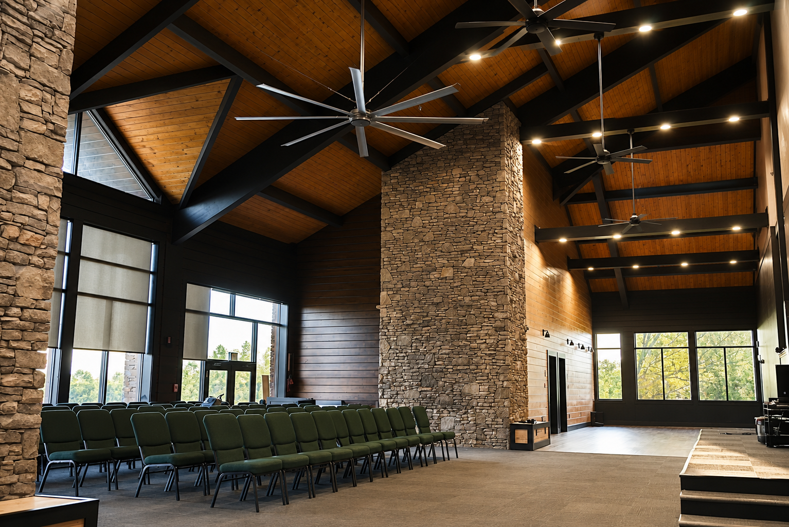 Interior of a large hall with a high wooden ceiling, black beams, prominent stone and wood walls, ceiling fans, and green chairs arranged in rows.