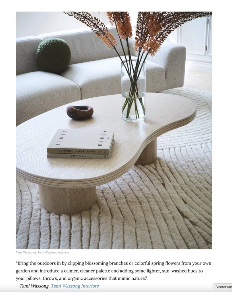 A minimalist living room with a beige, curved sofa, a light wooden kidney-shaped coffee table, a green round pillow, and a textured beige rug. On the table are decorative books, a small dark bowl, and a clear vase with tall dried flowers.