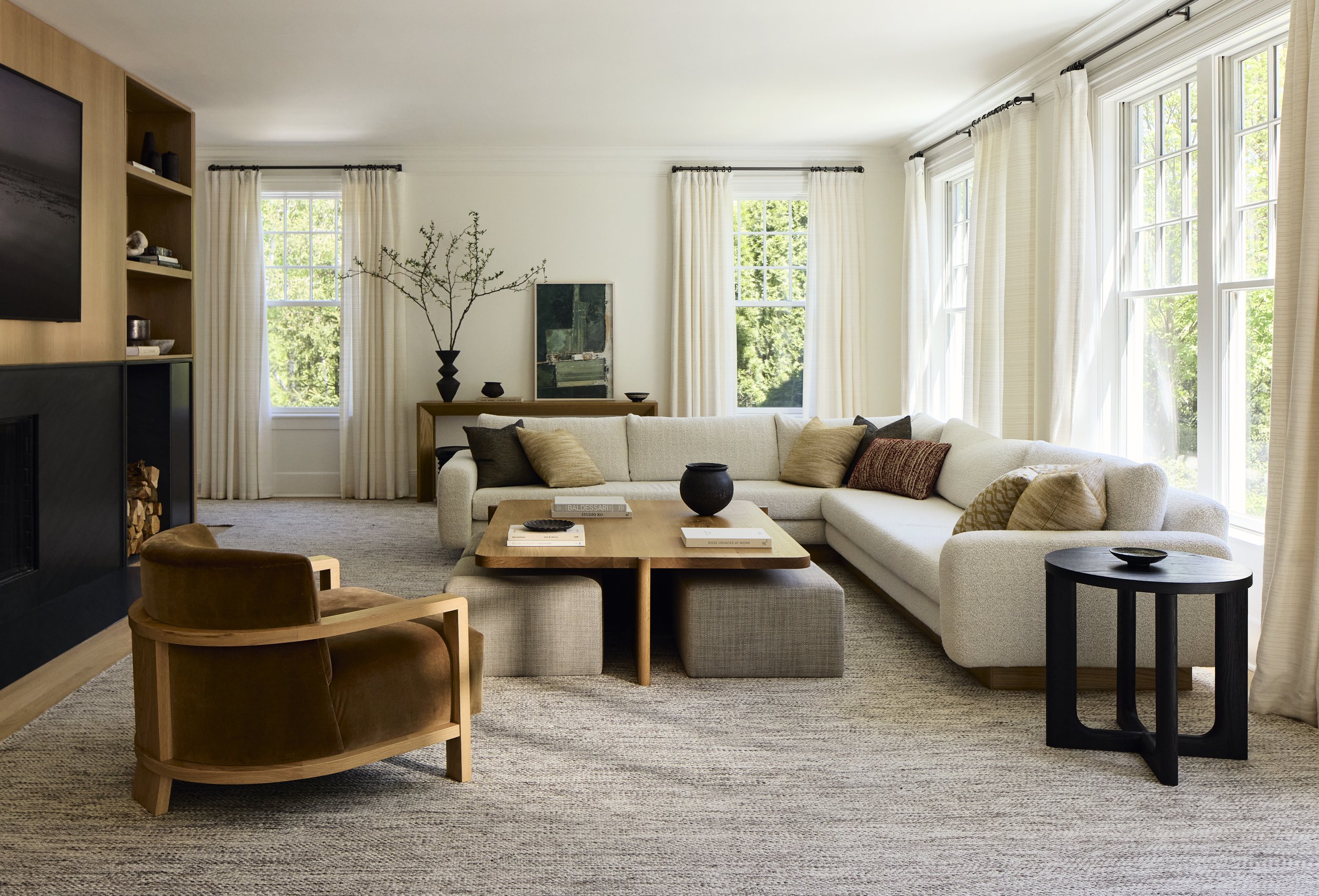 Modern living room with a large white sectional sofa, brown armchair, wooden coffee table, and side tables. Decor includes neutral cushions, art pieces, and a potted branch. Large windows with white curtains allow in ample natural light.