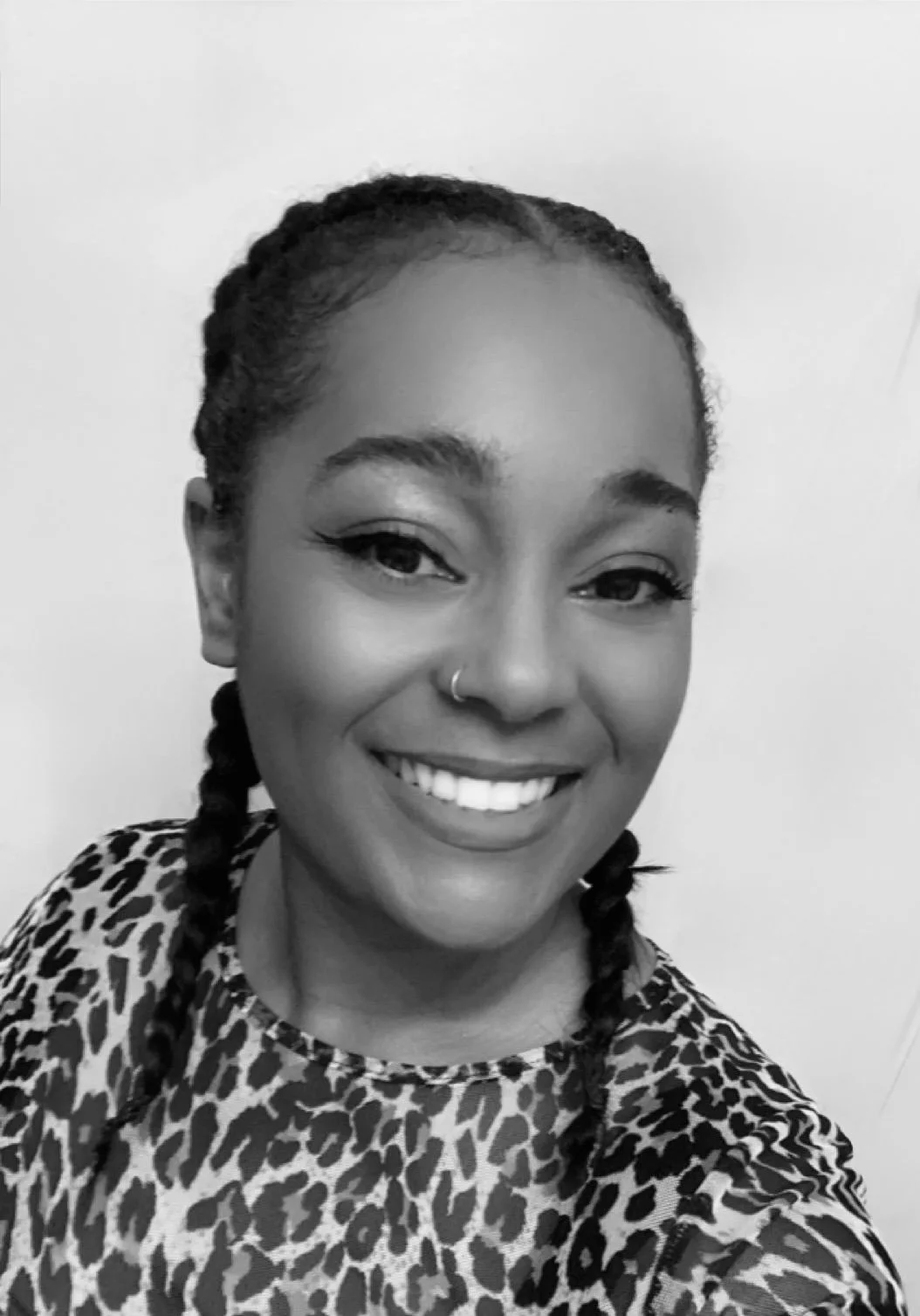 Black and white photo of a smiling woman with braided hair, wearing a leopard print top, with eyeliner and a nose ring.
