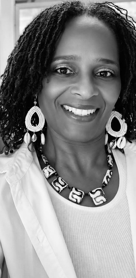 A smiling woman with short, curly hair wearing large, decorative earrings and a beaded necklace, dressed in a white shirt.