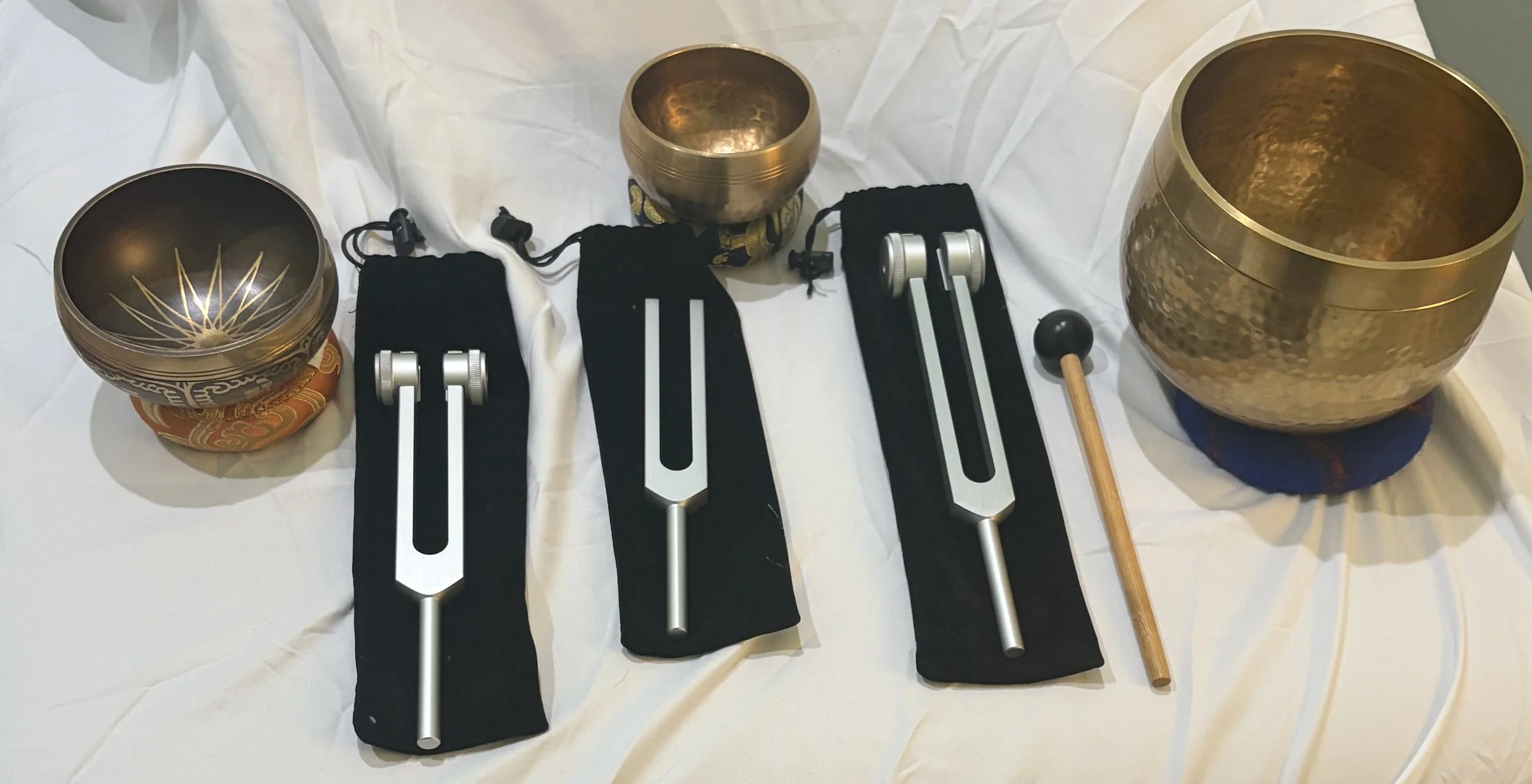 Set of three metallic singing bowls of varying sizes, each placed on a small cushion, along with three black mallets, metal tuning forks, and a wooden mallet with a black head, all arranged on a white fabric background.