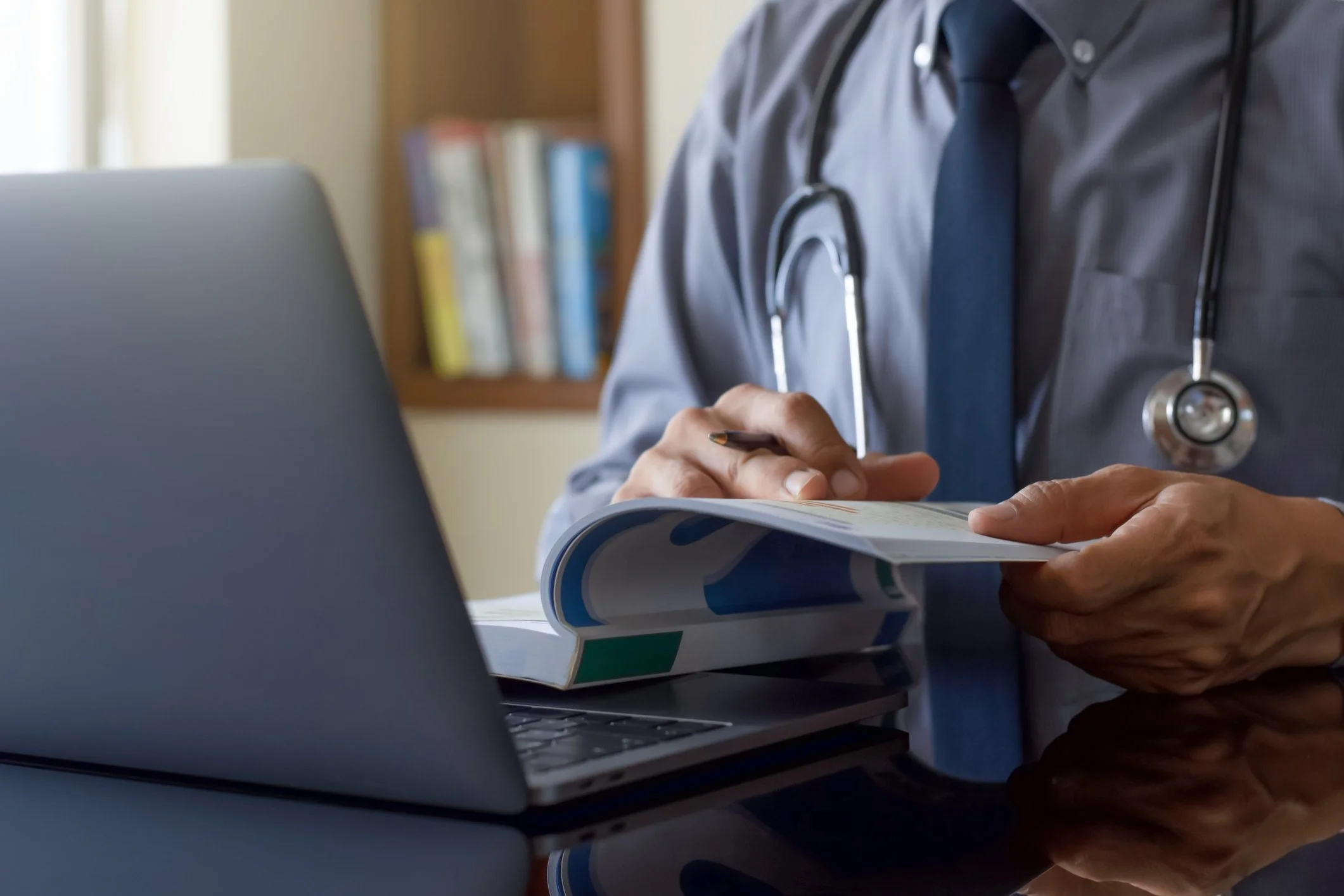 Physician reviewing clinical trial protocols and documents on a laptop with a stethoscope on the desk.
