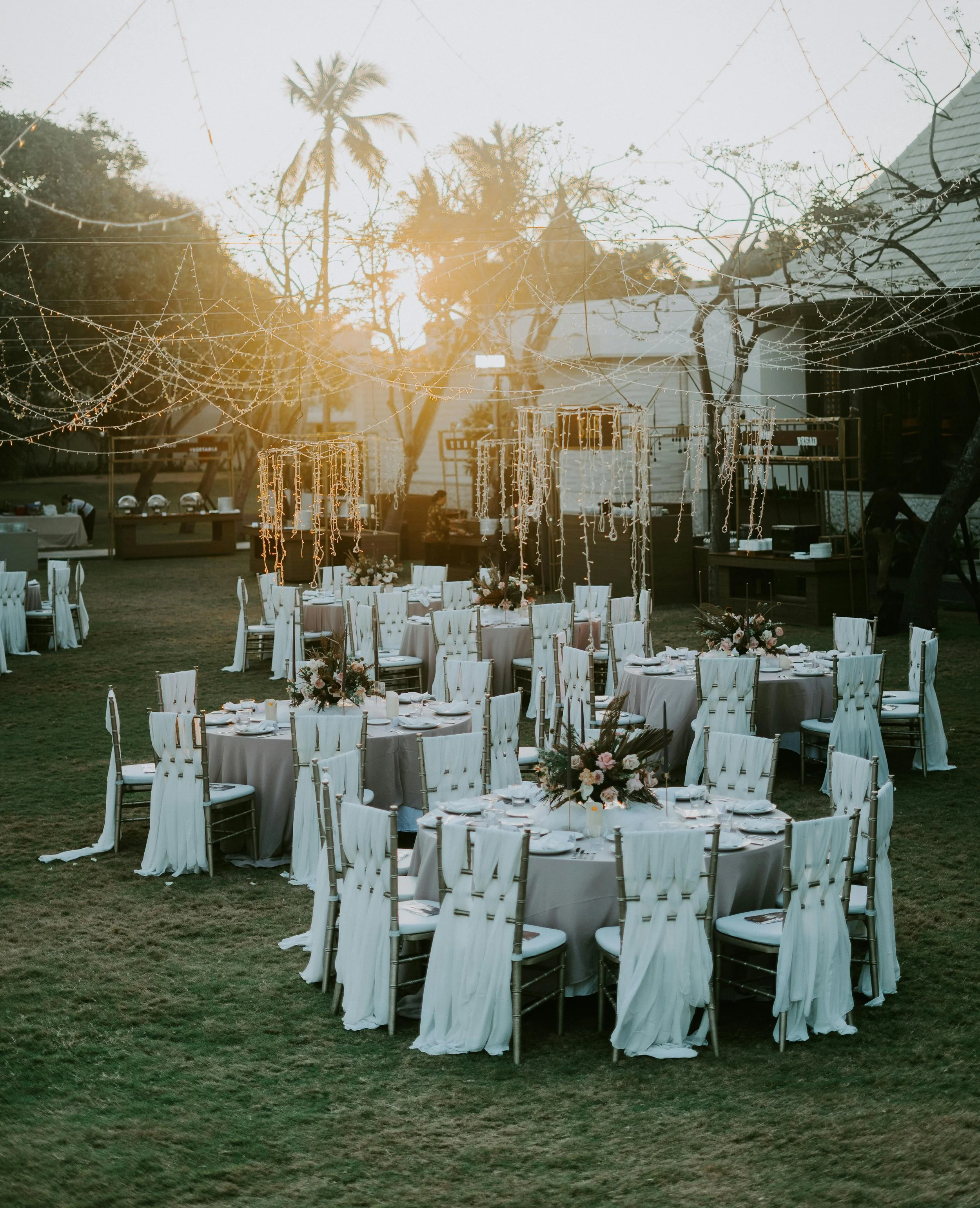 Outdoor wedding reception setup with round tables and white covered chairs, floral centerpieces, and hanging string lights at sunset.