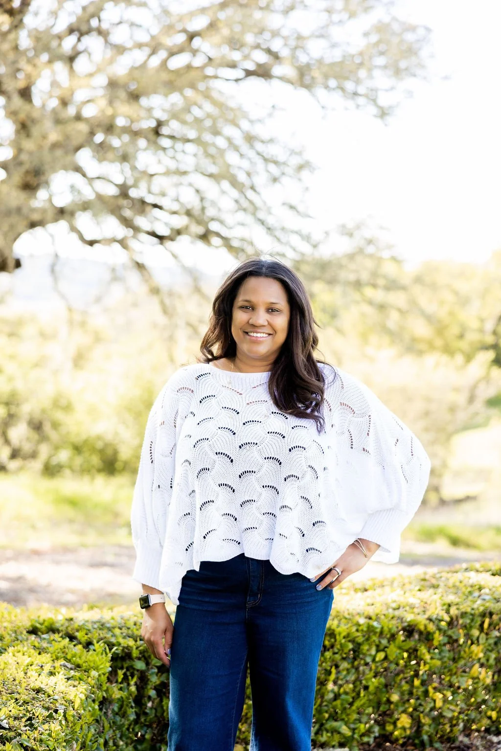 Woman in a white sweater and jeans standing outdoors with greenery and tree background.