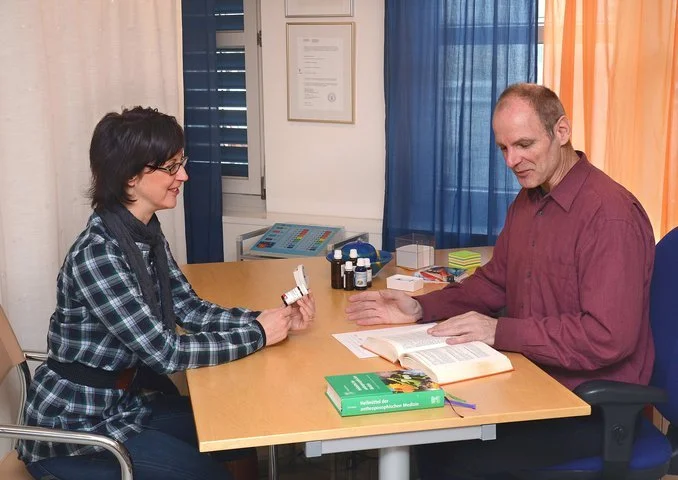 Zwei Menschen sitzen an einem Tisch in einem Raum, sprechen und schauen auf ihre Beobachtungen. Es sind Medikamente, eine offene Buch und ein geschlossener medizinischer Ratgeber auf dem Tisch.