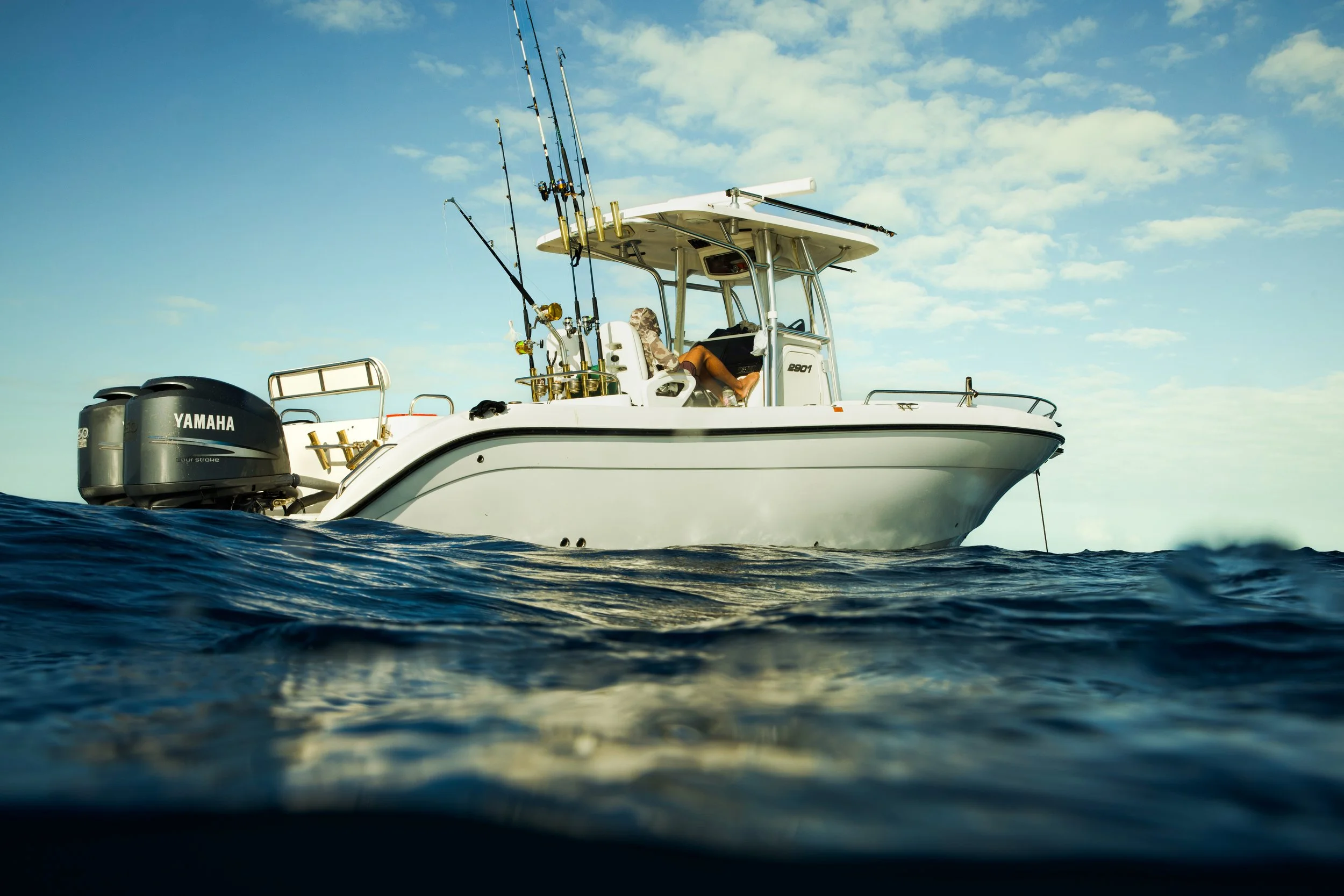 A white fishing boat with multiple fishing rods on the side, floating on the water under a partly cloudy sky.