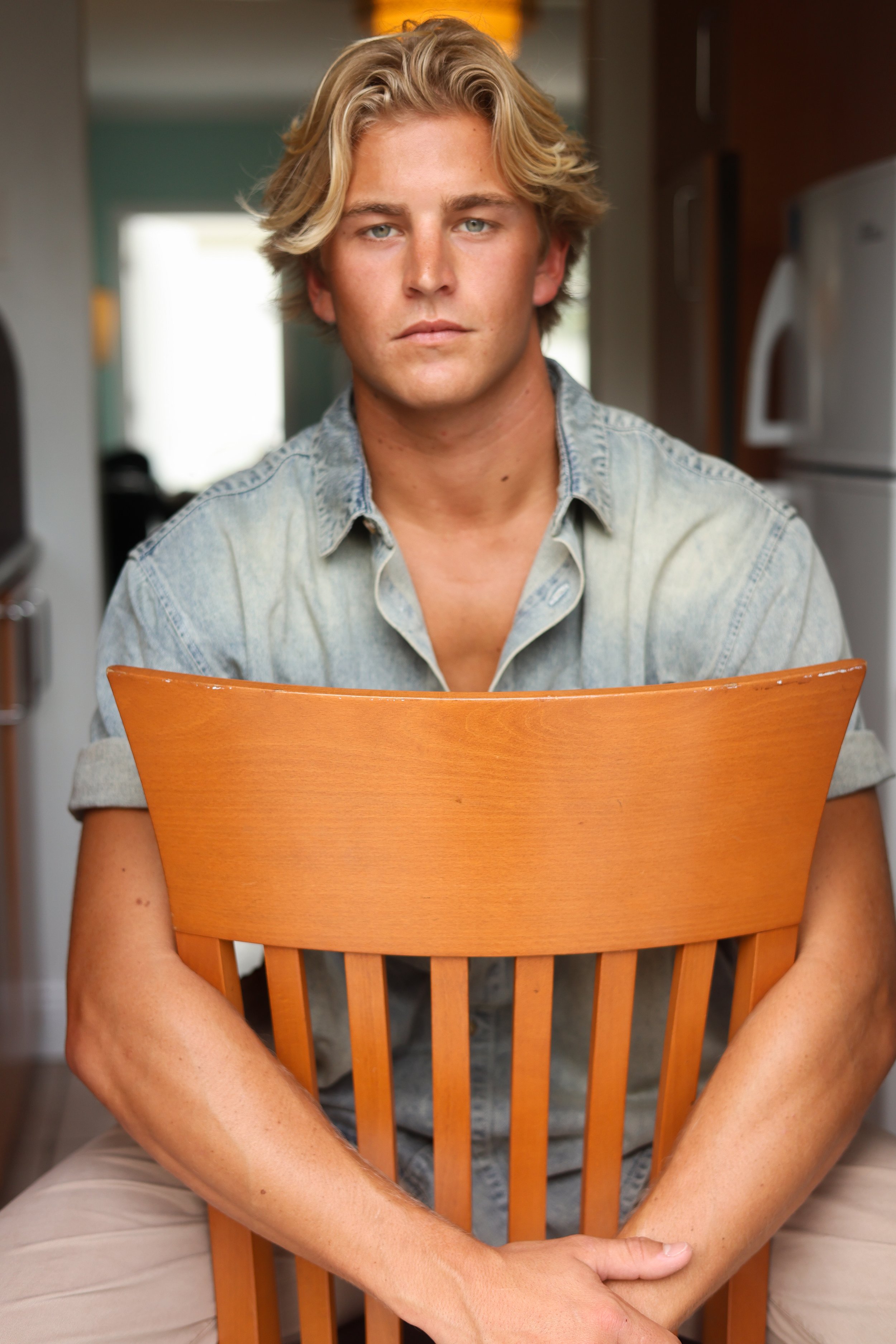 A young man with blond wavy hair wearing a light denim shirt, sitting behind an orange wooden chair, in a kitchen setting.