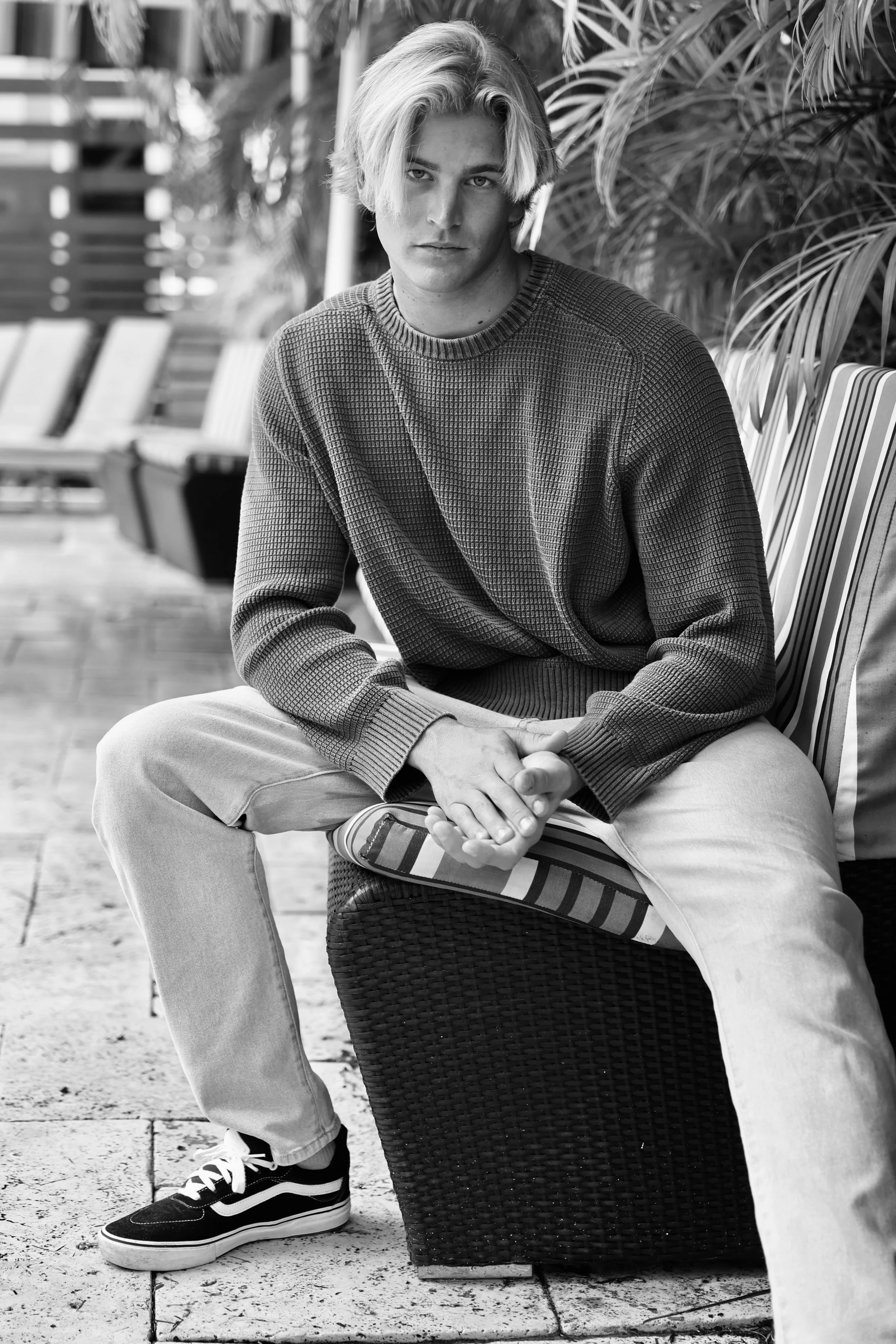 Black and white photo of a young man sitting outdoors on a wicker bench with striped cushion, wearing a textured sweater, light jeans, and sneakers, with palm plants in the background.