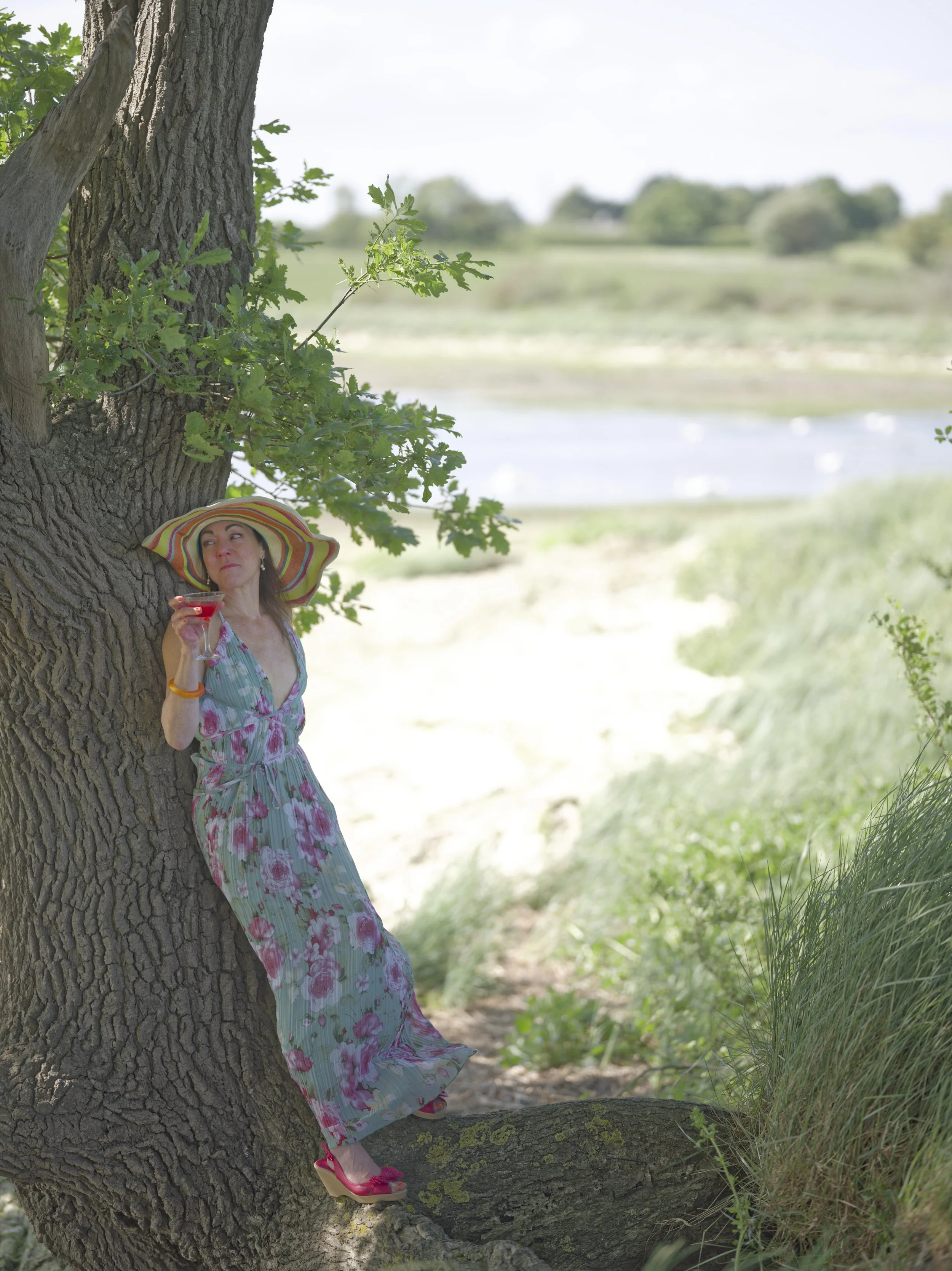 A woman in a floral dress and large sun hat leaning against a tree, holding a drink, in a natural outdoor setting with green grass, trees, and a body of water in the background.