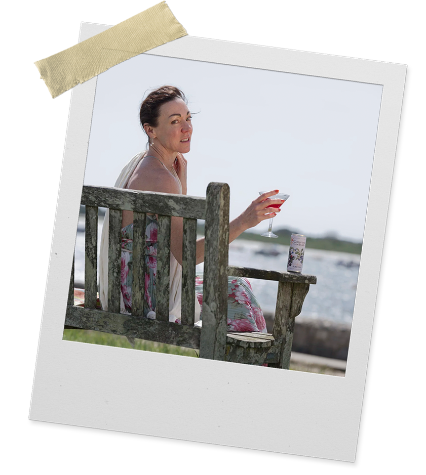 Woman sitting on a weathered wooden bench by the water, holding a wine glass, with a can of beverage on the bench, wearing a floral dress, outdoors on a sunny day.