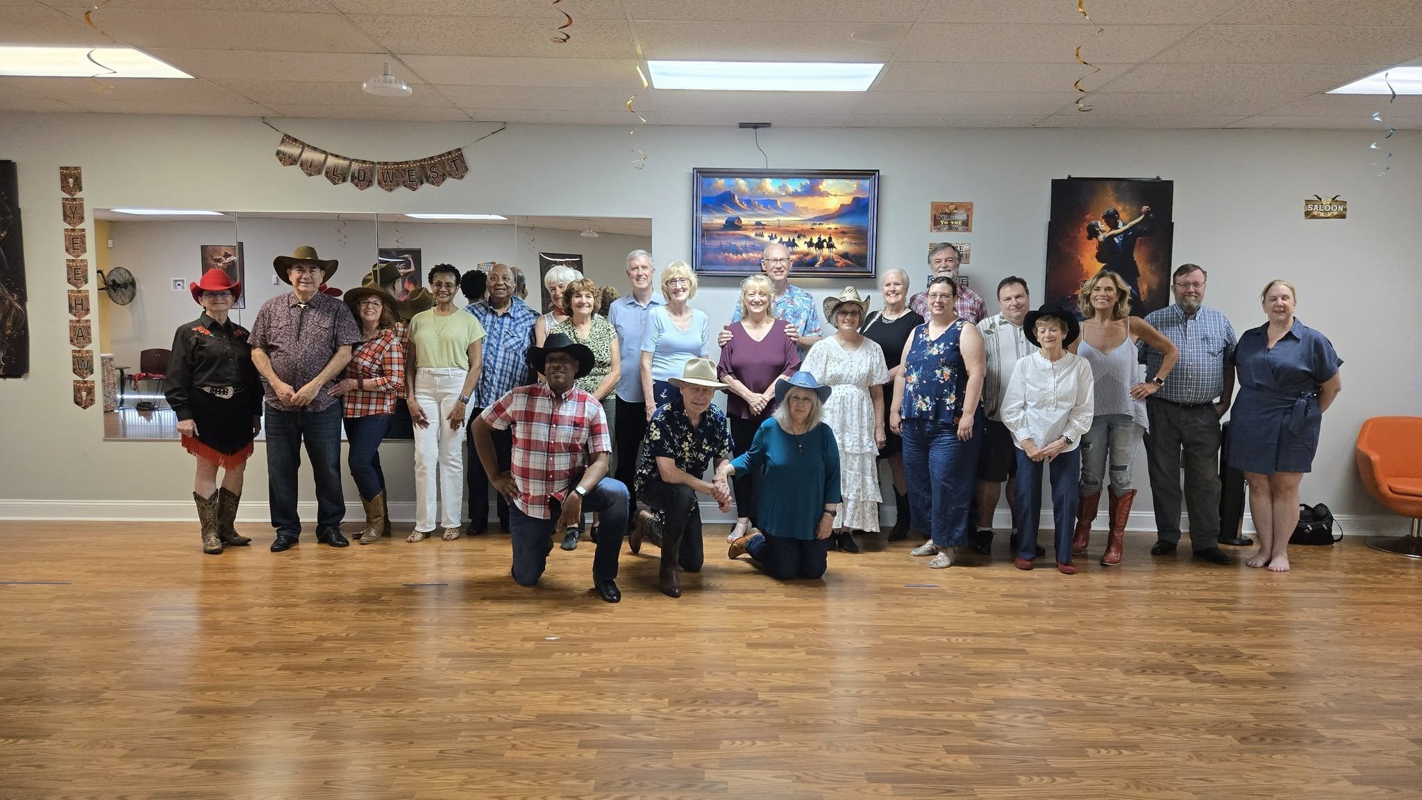 Group of people dressed in cowboy and Western-themed attire posing indoors for a photo, with framed artwork and decorations on the wall.