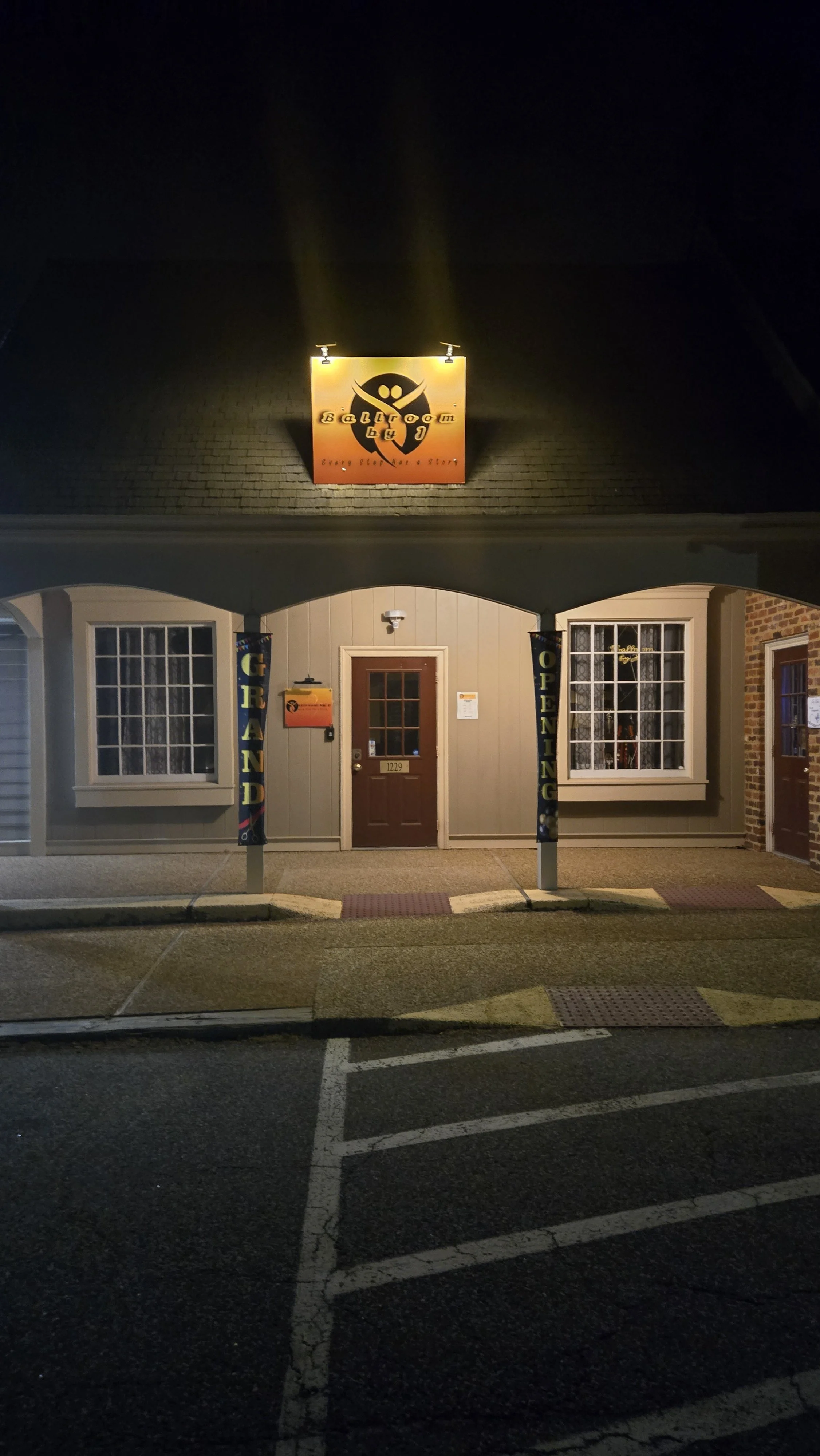 Nighttime view of the front entrance of a restaurant called 'Balroom by'. The sign above the entrance is illuminated, showing a logo with a smiling face and text. There are two large windows on either side of the door, and banners hanging from poles that say 'GRAND OPENING'. The building has beige siding and a small porch area.