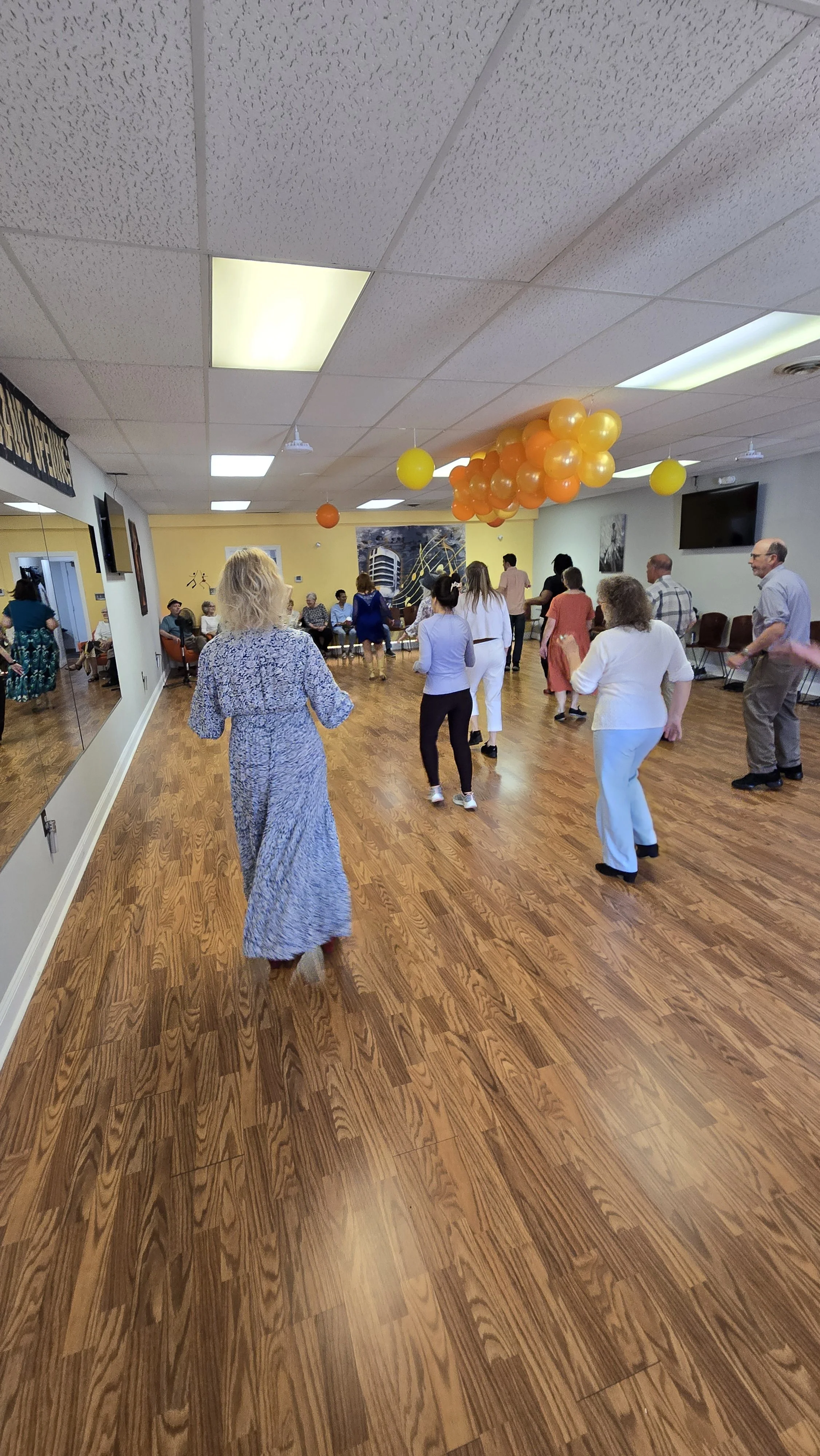 People dancing in a room decorated with orange and yellow balloons, with a wall featuring artwork and a large mirror.
