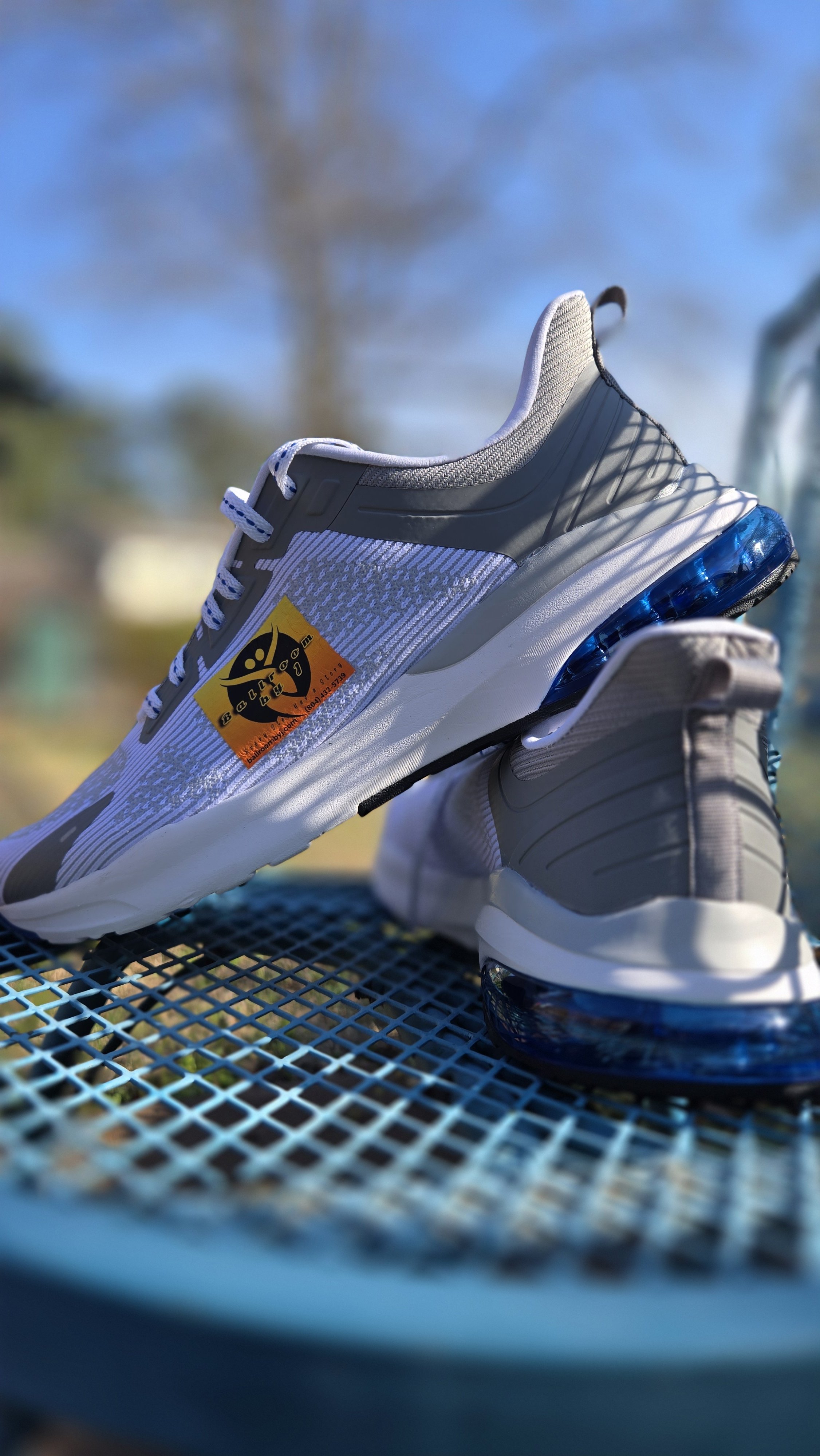 Close-up of clear Nike Air Max sneakers with a black and orange emergency sticker on the side, placed on a blue metal table outdoors with a blurred background of trees and sky.