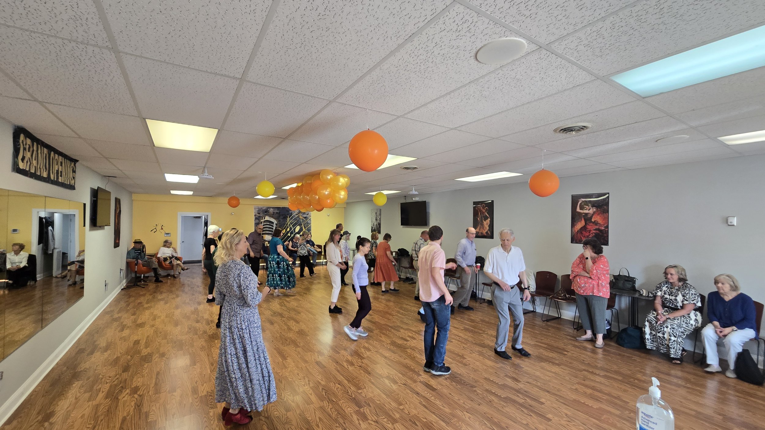 People gathered in a spacious room with wooden floors, decorated with orange and yellow balloons, participating in a social event or gathering.