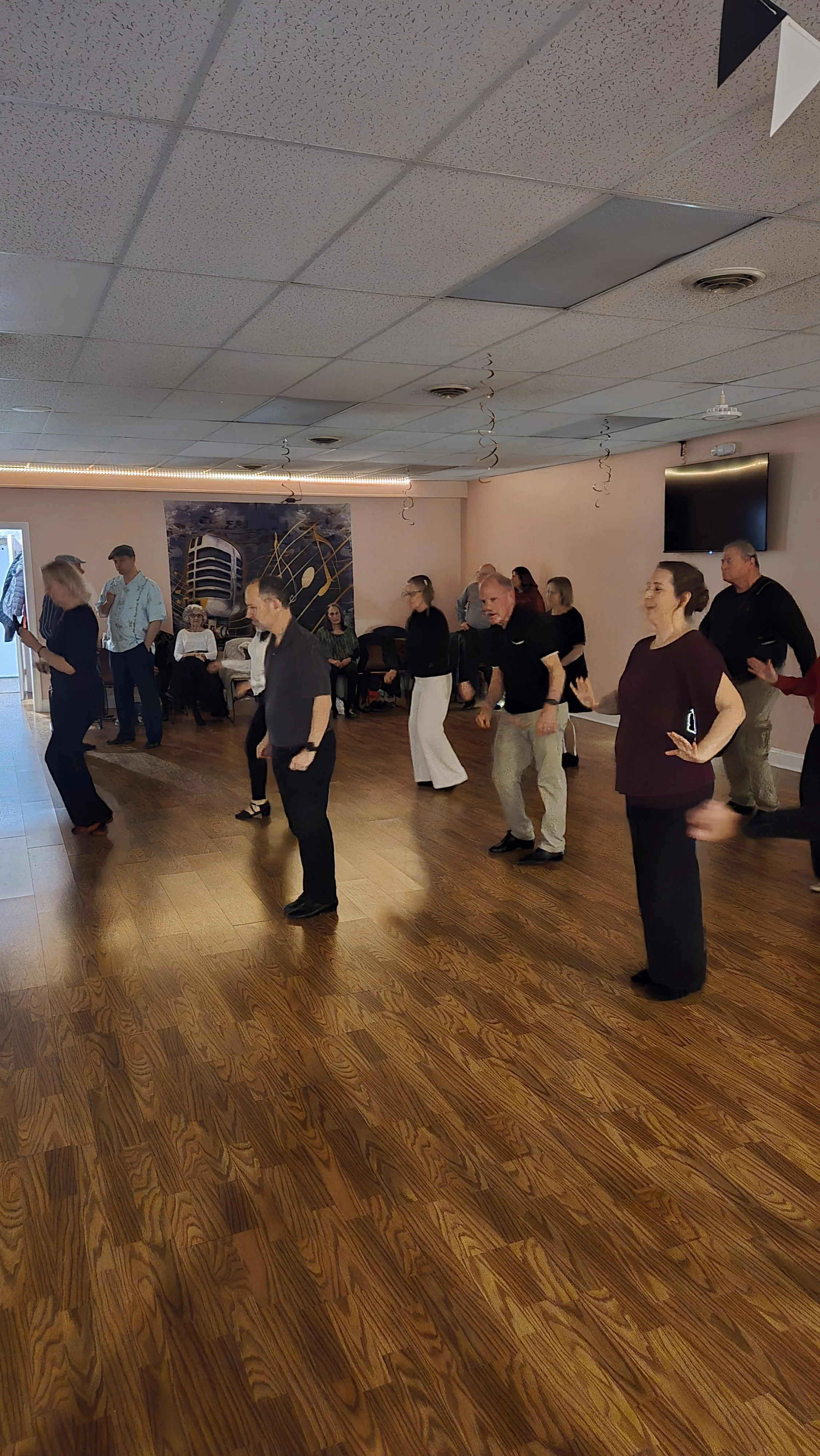 People dancing in a room with wood flooring, decorated with black and white flags hanging from the ceiling. Seating area with some individuals seated and a large wall art with a microphone in the background.