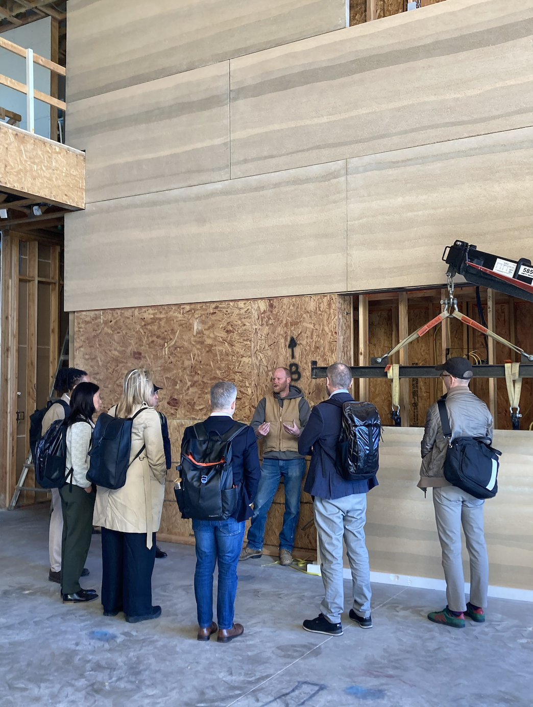 A group of people listening to a speaker inside a construction site, with rammed earth wall. 