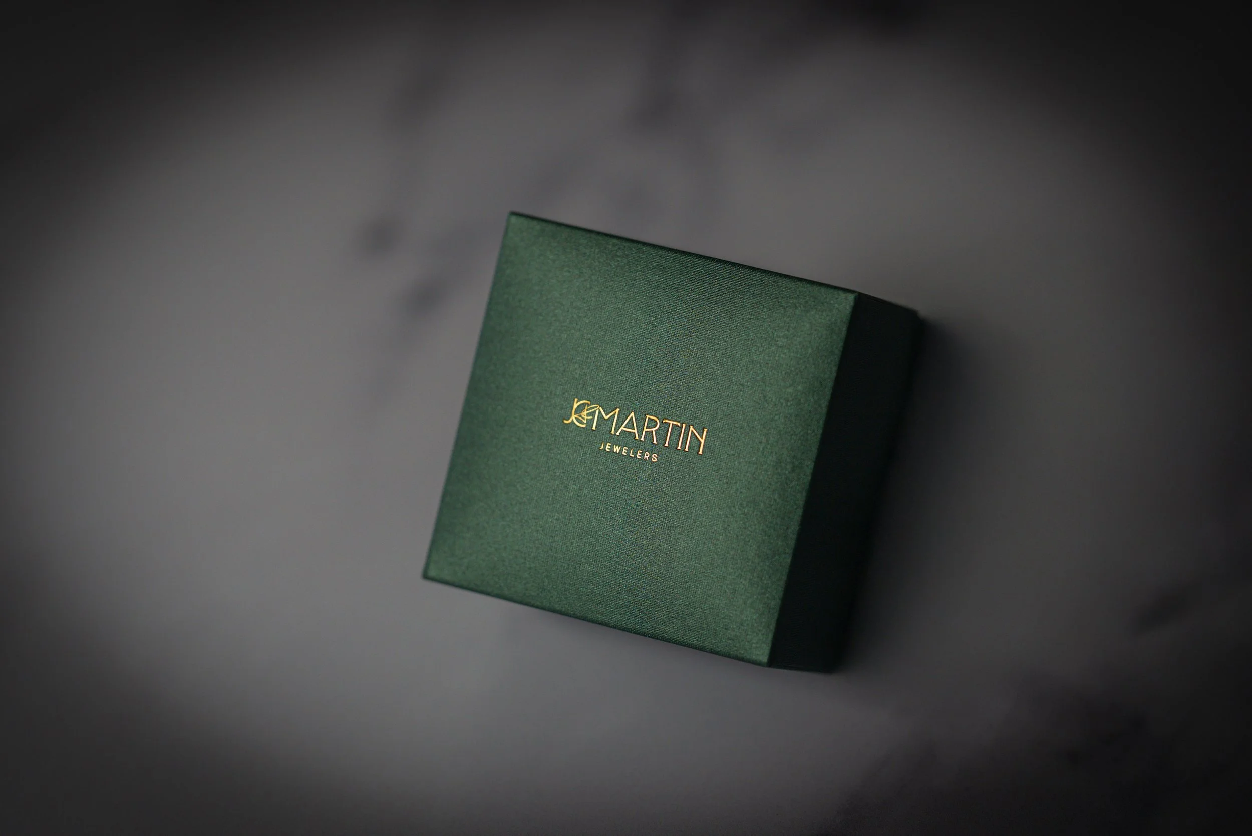 A green jewelry box on marble slab featuring the logo of JC Martin Jewelers