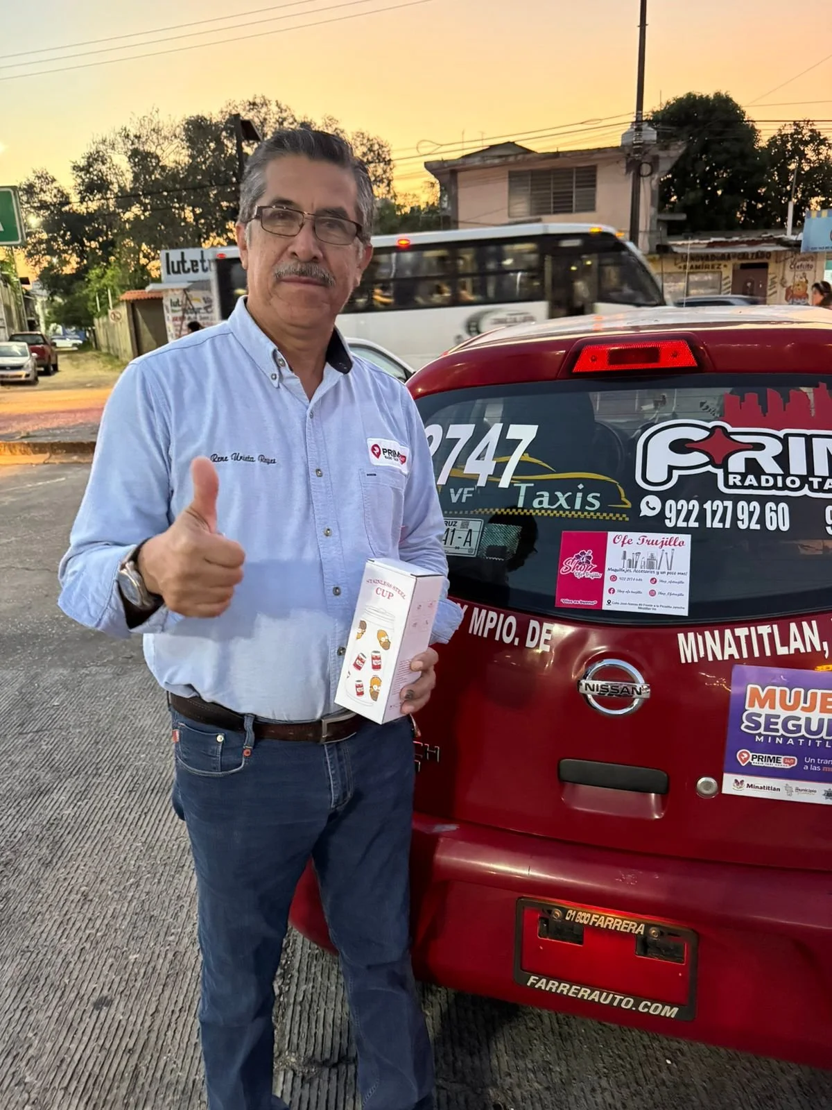 Hombre de cabello y bigote gris, con gafas, vestido con camisa azul con logotipo de PRD, sosteniendo una caja de dulces o snacks, y dando pulgar hacia arriba, de pie junto a un vehículo de taxi en una calle urbana al atardecer.