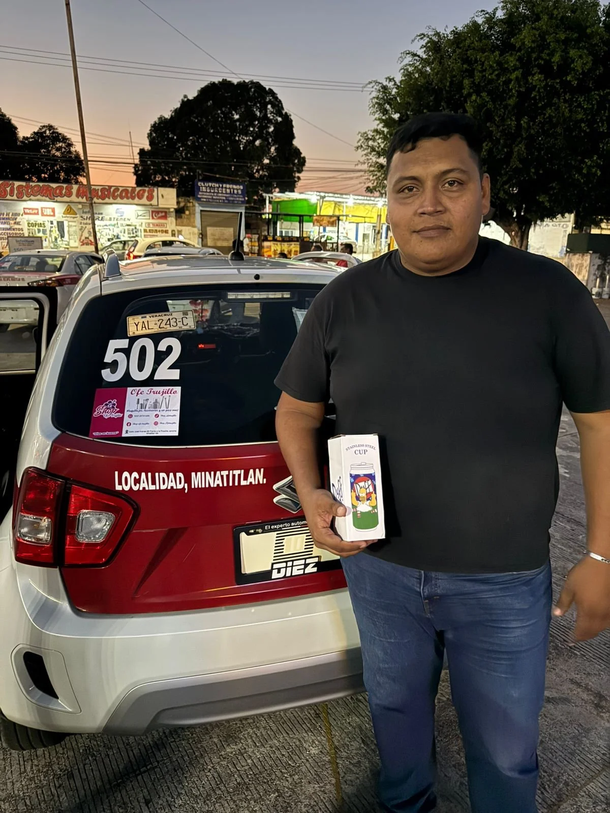 Un hombre de tez morena, con camiseta negra y jeans, sostiene una caja de taza de acero inoxidable con un diseño colorido y un logo, parando frente a un coche con placa de Veracruz, en una calle al atardecer, con tiendas, árboles y otros vehículos en