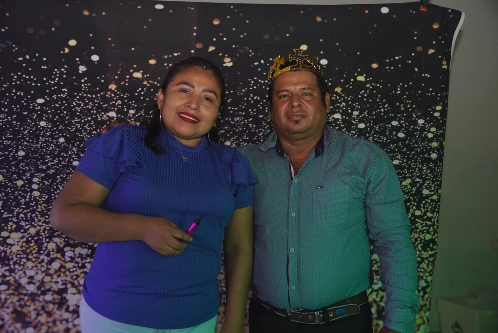 Dos personas de pie frente a un fondo con puntos blancos y naranjas sobre fondo negro, la mujer lleva blusa azul y la corona en la cabeza del hombre indica que está celebrando un cumpleaños o evento especial.