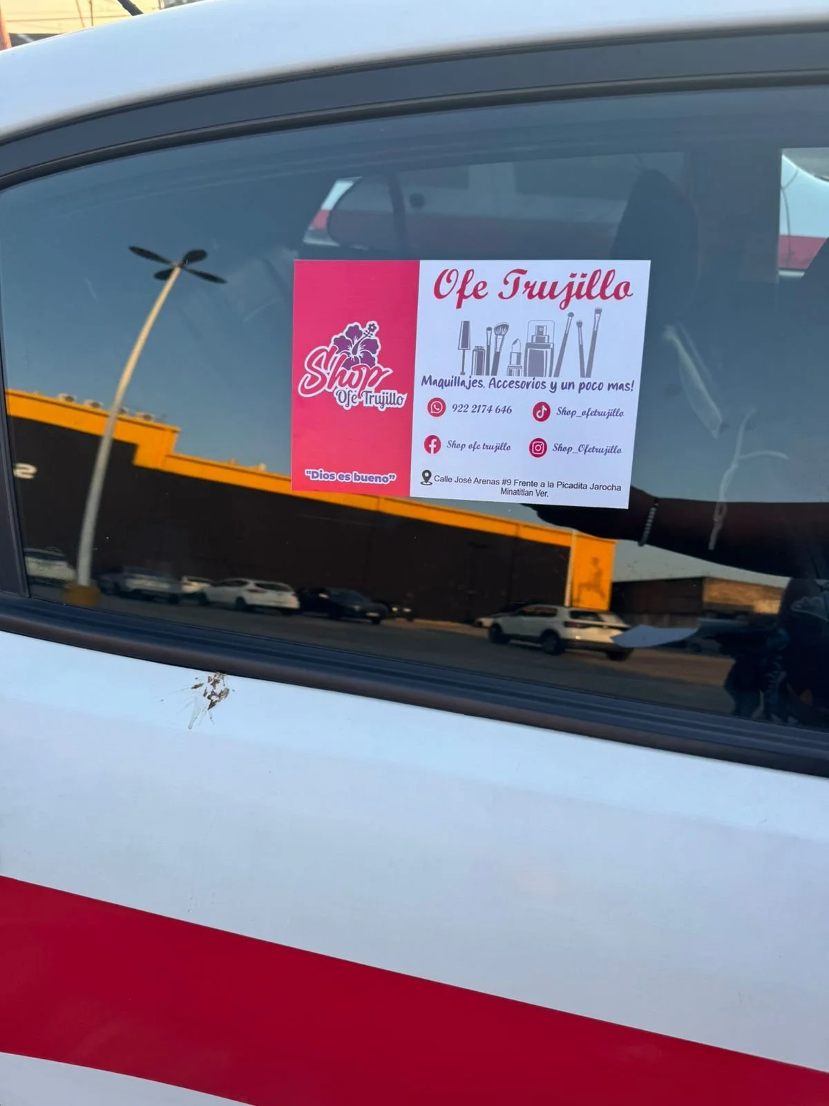 Un cartel en el parabrisas de un auto que promociona un negocio llamado Shop ofe Trujillo, que ofrece maquillaje, accesorios y otros servicios. La dirección y redes sociales también están en el cartel.
