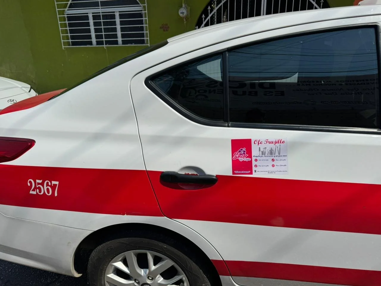 Automóvil blanco con franja roja y el número 2567, con cartel de venta o anuncios en la ventana, en un entorno urbano con muro verde y ventana con rejas.
