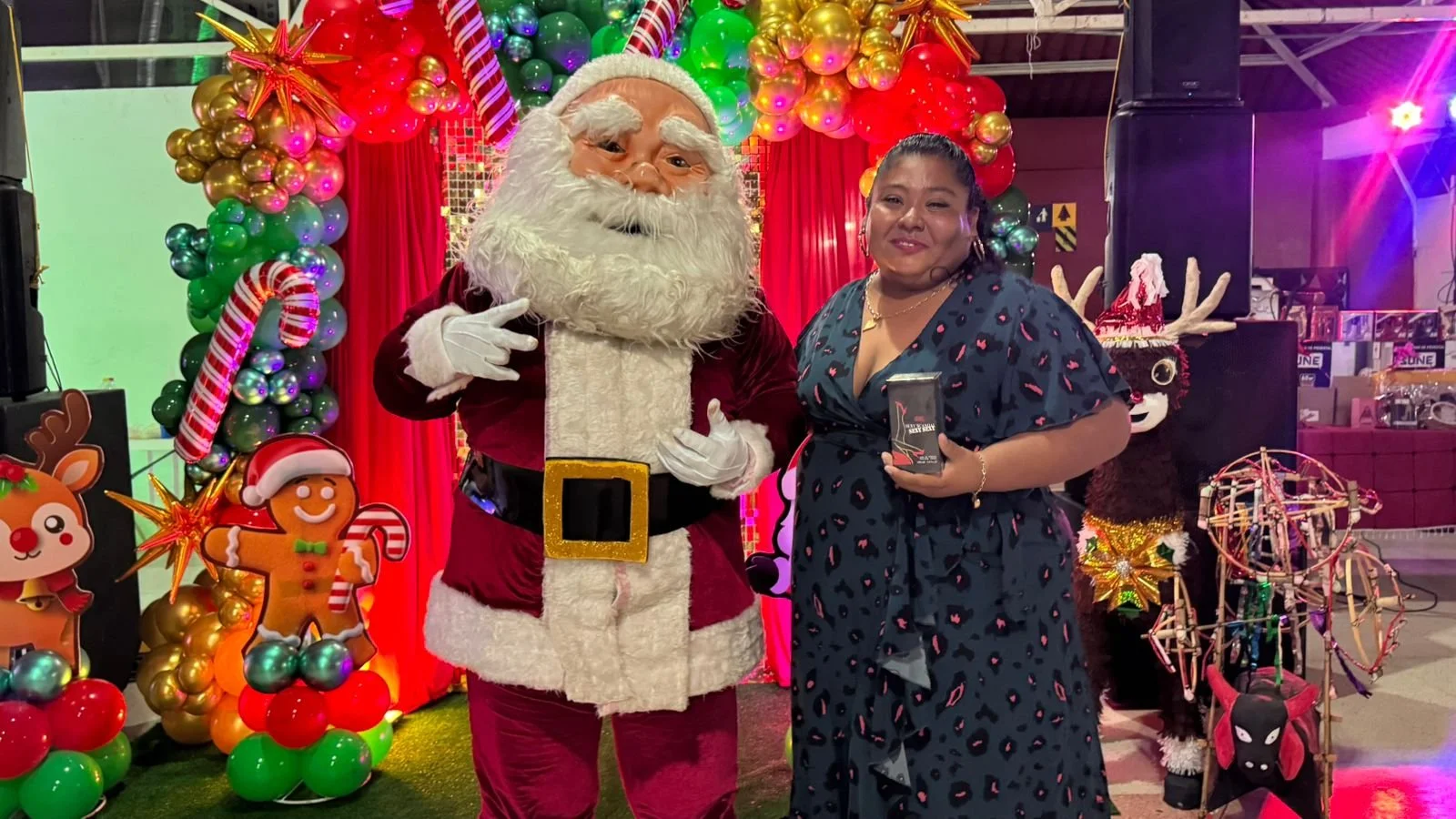 Una mujer y Santa Claus posando en un escenario navideño decorado con globos, guirnaldas de colores y figuras de jengibre, en un ambiente festivo.