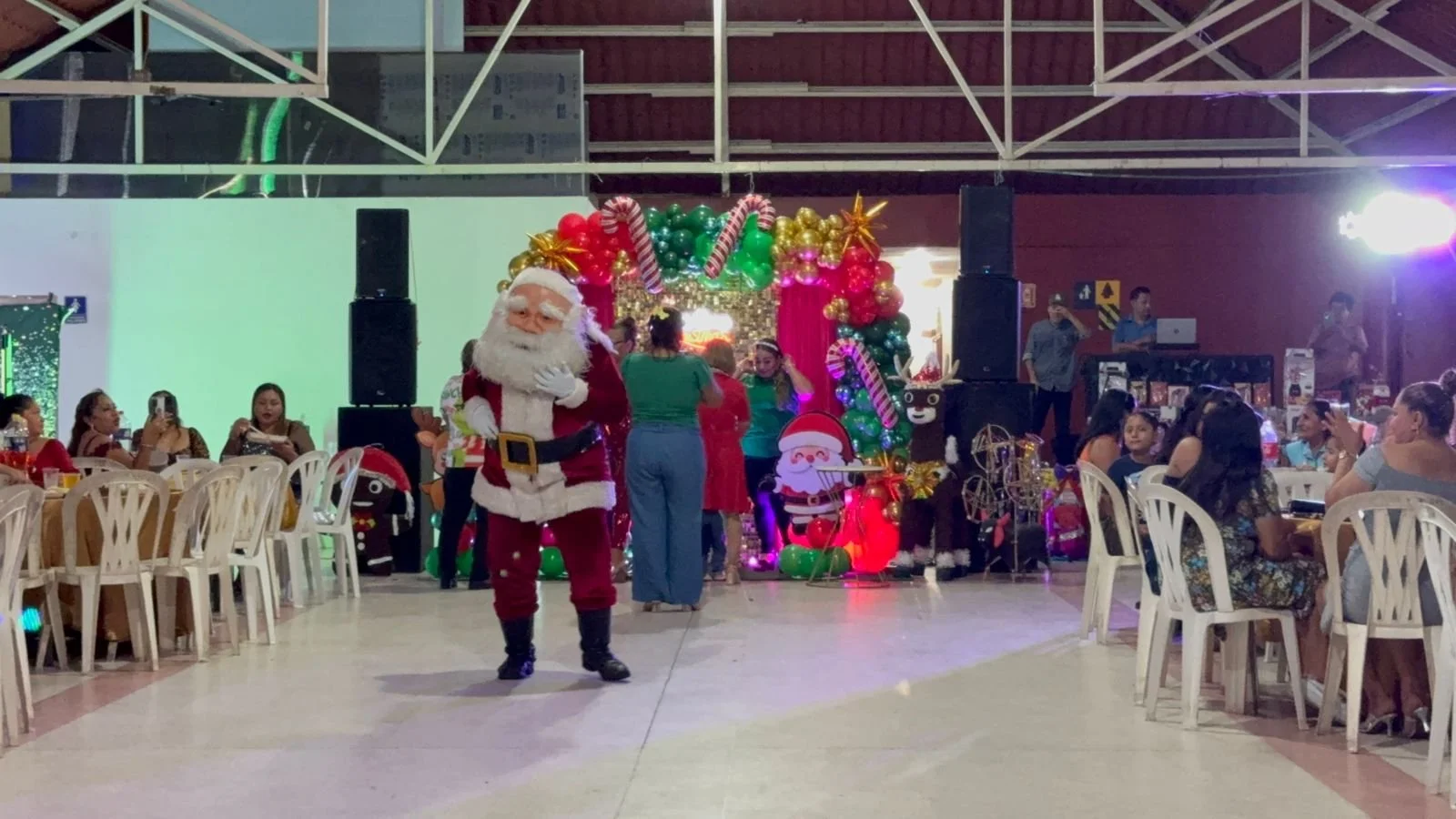 Hombre disfrazado de Santa Claus en un evento navideño con decoraciones festivas y un fondo de globos y personajes de Navidad.