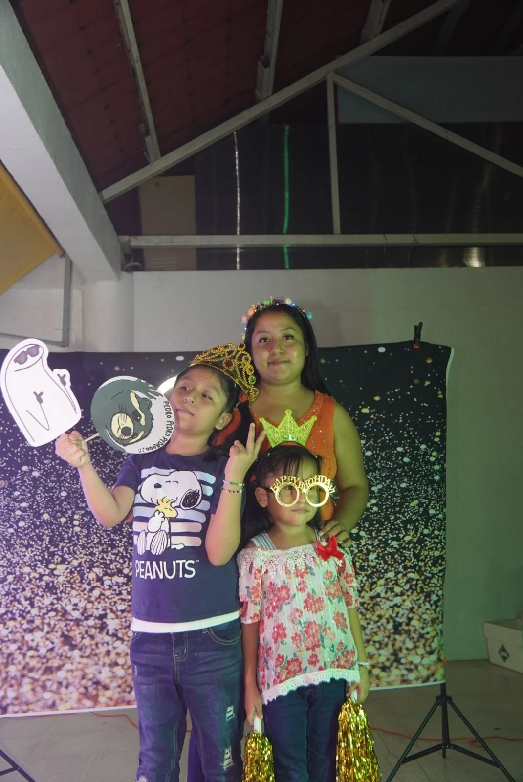 Celebración de cumpleaños infantil con tres niños y una mujer posando frente a un fondo decorativo, usando accesorios divertidos y disfraces. Uno de los niños sostiene un cartel y una máscara, otra niña lleva gafas de cumpleaños y una corona, la muje