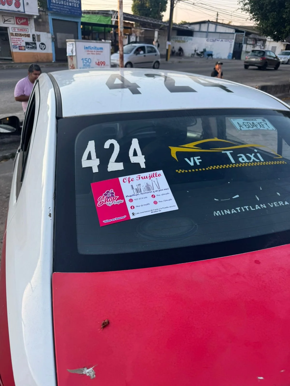 Vista de la parte superior de un taxi con varias pegatinas y números, incluyendo el número 424 en la parte trasera y el logotipo VF Taxi, en una calle con personas y vehículos de fondo.