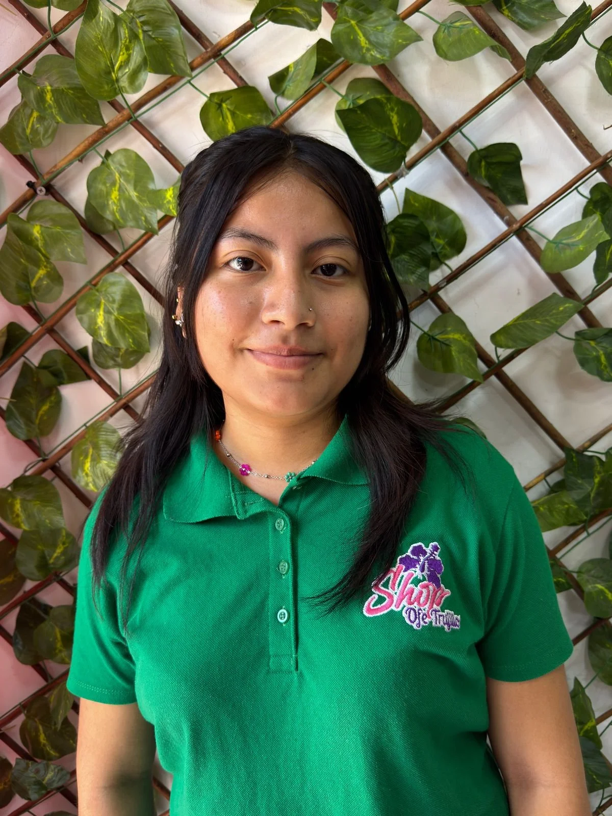 Una joven vestida con una camiseta verde con un logotipo en el lado izquierdo que dice 'Shop Oje Trujillo', con cabello oscuro y largo, usando aretes pequeños y un collar de bolitas de colores, de pie frente a una pared decorada con enredaderas verde