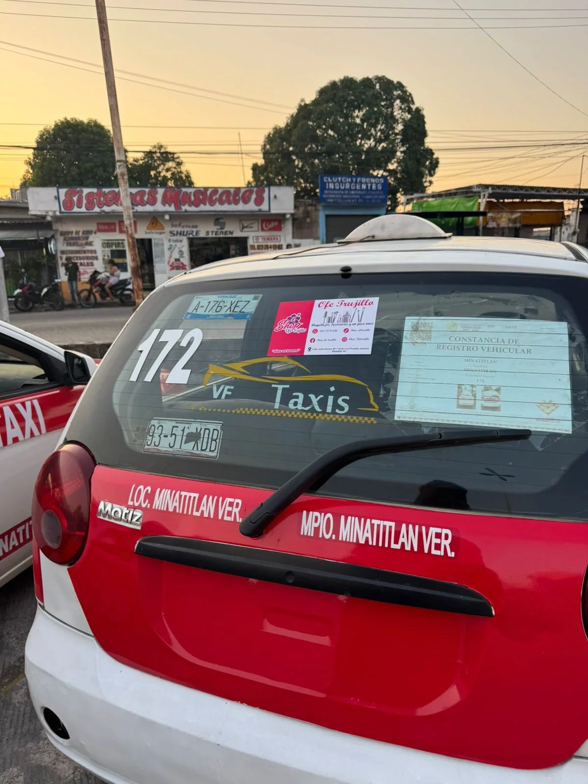 Parte trasera de un coche de taxi con varias pegatinas y papeles en la ventana. En el fondo, un establecimiento con un cartel que dice 'Sistemas Musicales' y una calle con motos y árboles.