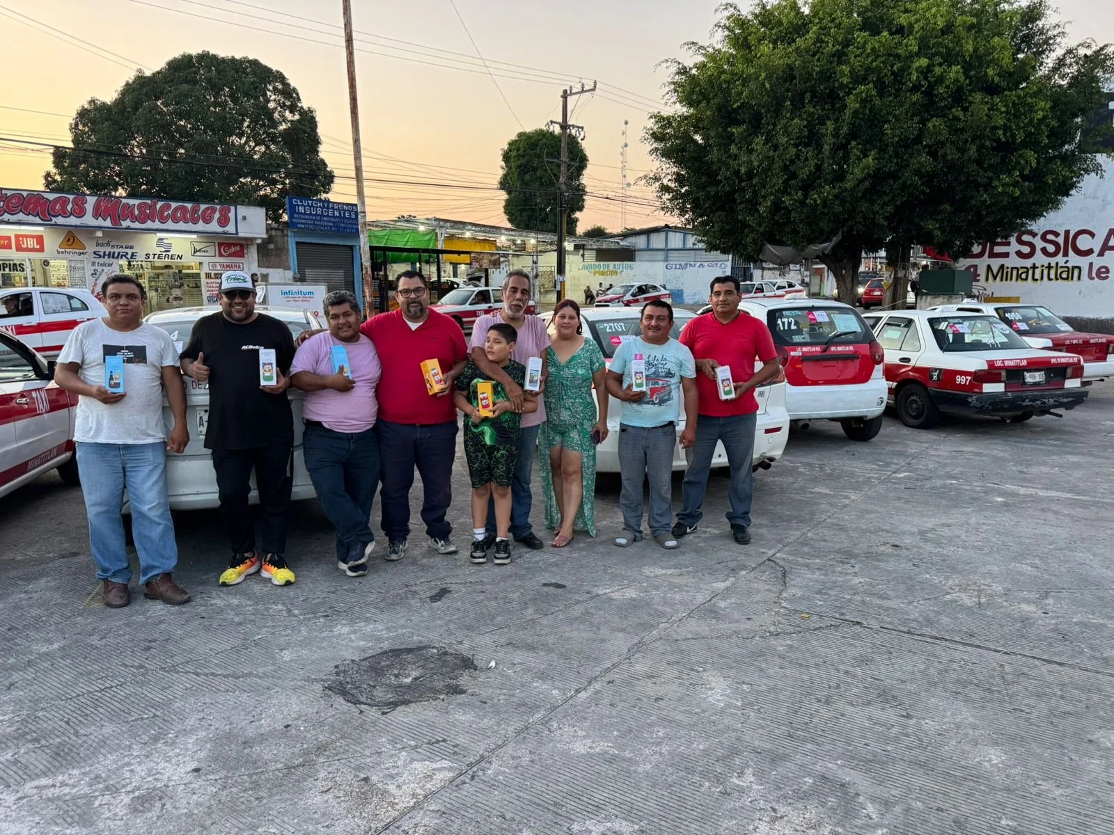 Grupo de ocho personas de diferentes edades y géneros, algunos con camisetas rojas, sosteniendo productos en frente de varios autos de taxi en un estacionamiento. Fondo con árboles, tiendas y postes eléctricos al atardecer.