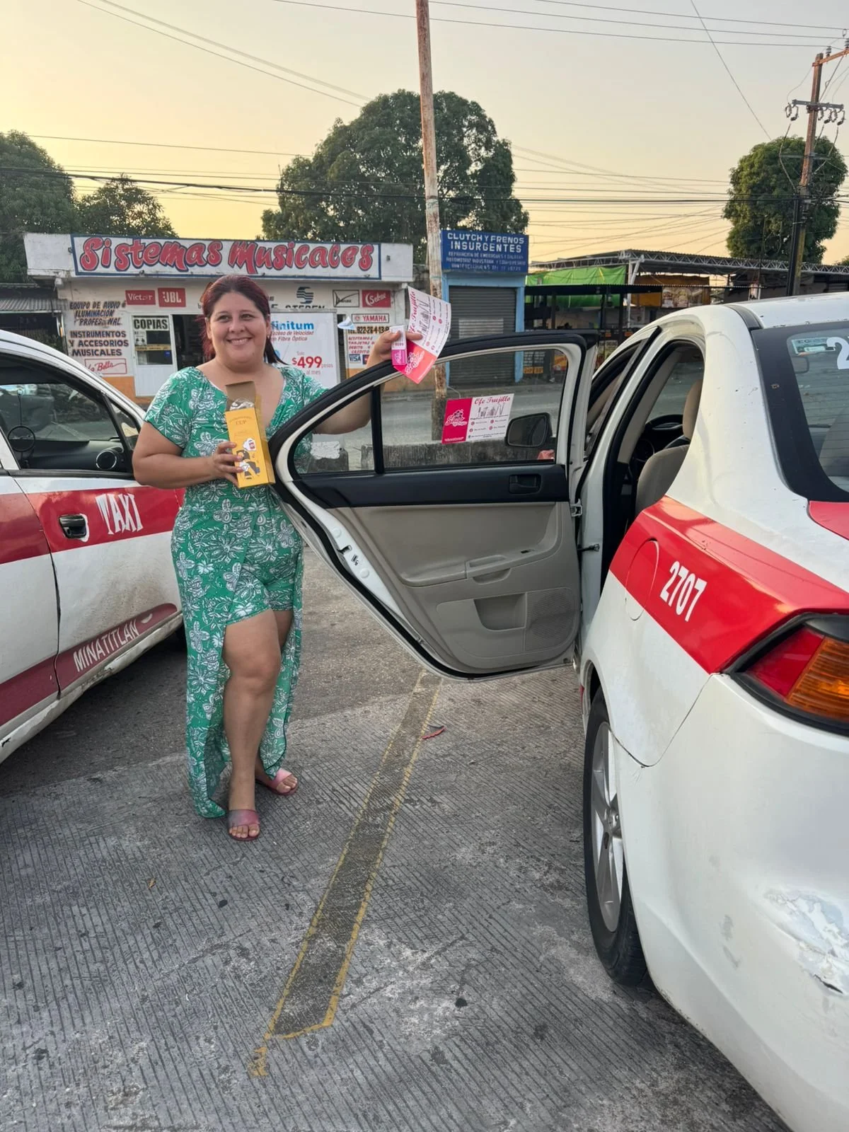 Una mujer de cabello castaño en un conjunto verde con flores, sosteniendo un producto en una mano y algunos folletos en la otra, junto a un taxi blanco con línea roja en un estacionamiento al aire libre.