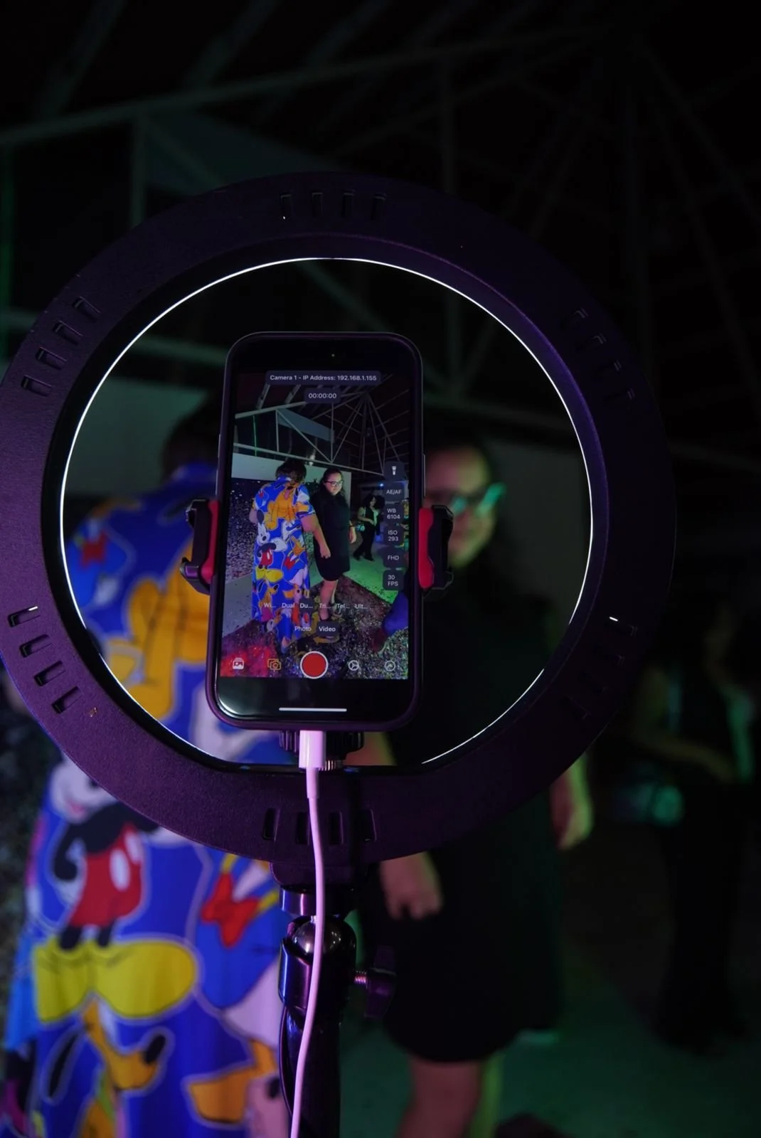 Vista a través del visor de un teléfono grabando a dos personas en un escenario con fondo oscuro y estructura metálica, rodeada de un aro de luz