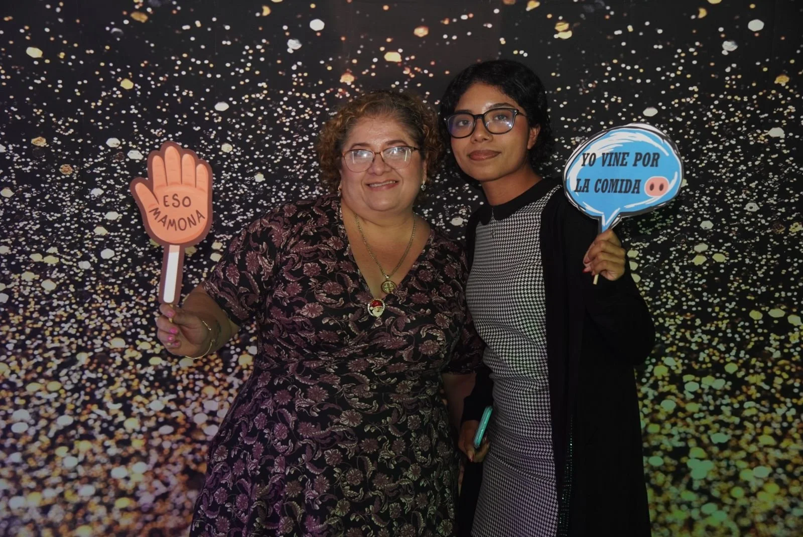 Dos mujeres posando juntas frente a un fondo de confetti. La mujer a la izquierda sostiene un cartel que dice 'Eso mamona' y la mujer a la derecha tiene un cartel que dice 'Yo vine por la comida'.