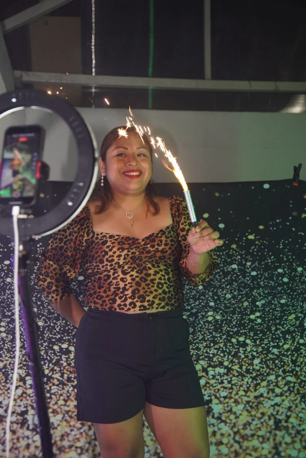 Mujer sonriendo sosteniendo una bengala encendida, usando una blusa de estampado leopardo y pantalones cortos negros, con un fondo lleno de confeti y una cámara en primer plano.