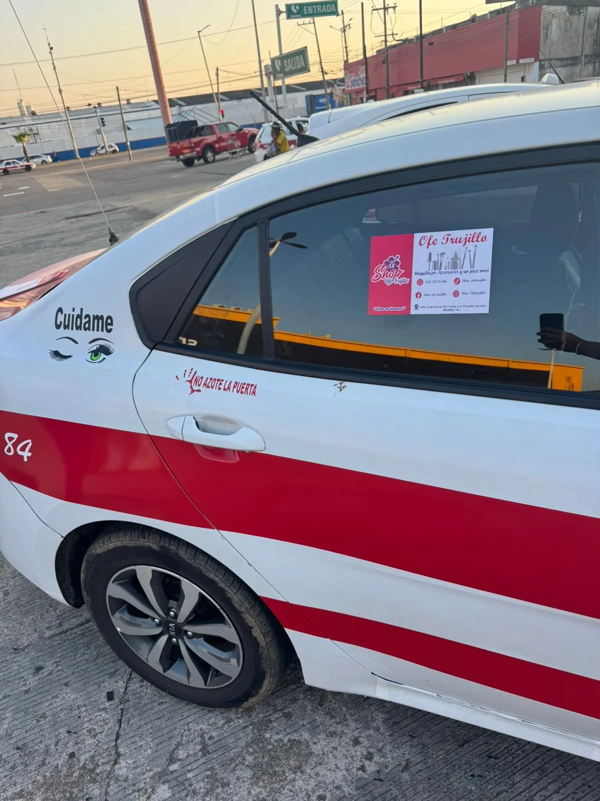 Automóvil con señal de advertencia que dice 'No azote la puerta', y un cartel en la ventana con información del comercio 'Ofe Trujillo', en un estacionamiento abierto.