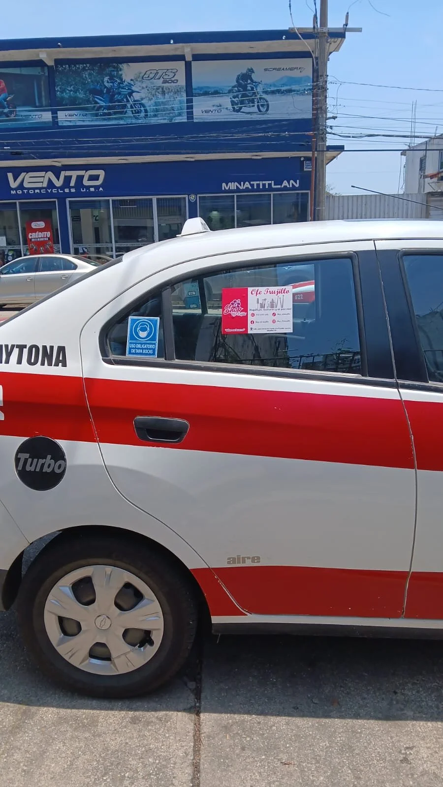 Auto de policía con un aviso en la ventana que promociona una cafetería llamada 'Ofe Trujillo' y una tienda de motocicletas llamada 'Vento'.