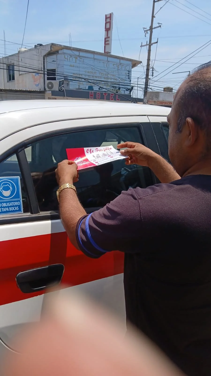 Un hombre coloca una hoja en un automóvil, con un cartel de '¡Feliz Inauguración'.