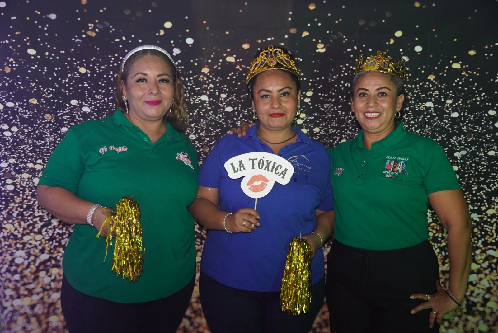 Tres mujeres con corona y camisetas de colores, sosteniendo pompones dorados y un cartel que dice 'La Tóxica' con boca annak en medio, en un fondo con luces brillantes.