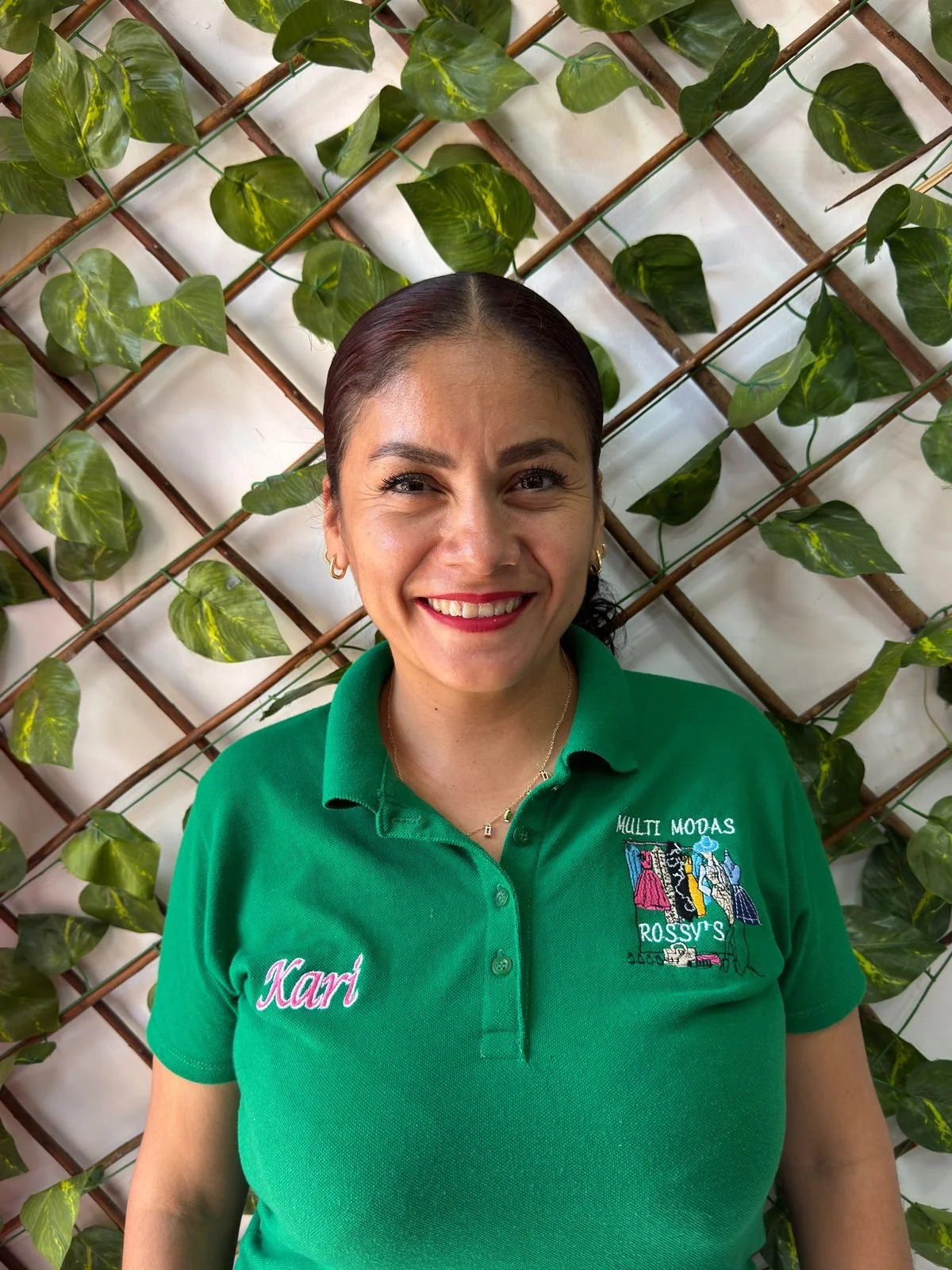 Mujer sonriendo con uniforme verde que tiene el nombre Karí y el logotipo de una tienda de ropa llamada Multi Modas Rossy's, posando frente a una pared decorada con enredadera de hojas verdes.