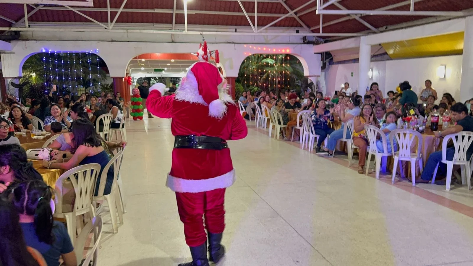 Persona disfrazada de Santa Claus caminando en un evento con muchas personas sentadas en mesas decoradas para celebrar la Navidad.