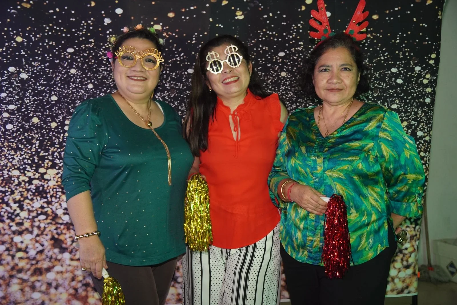 Tres mujeres con accesorios festivos en una celebración de fin de año, con fondo de confeti dorado y negro.