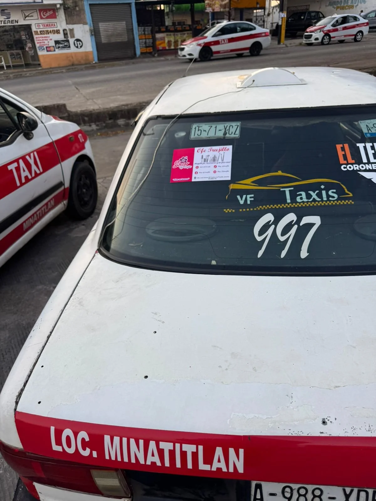Taxi blanco con letrero en la parte trasera que dice 'LOC. MINATITLÁN', con anuncios y pegatinas en la parte trasera, en una calle con otros taxis y comercios en el fondo.