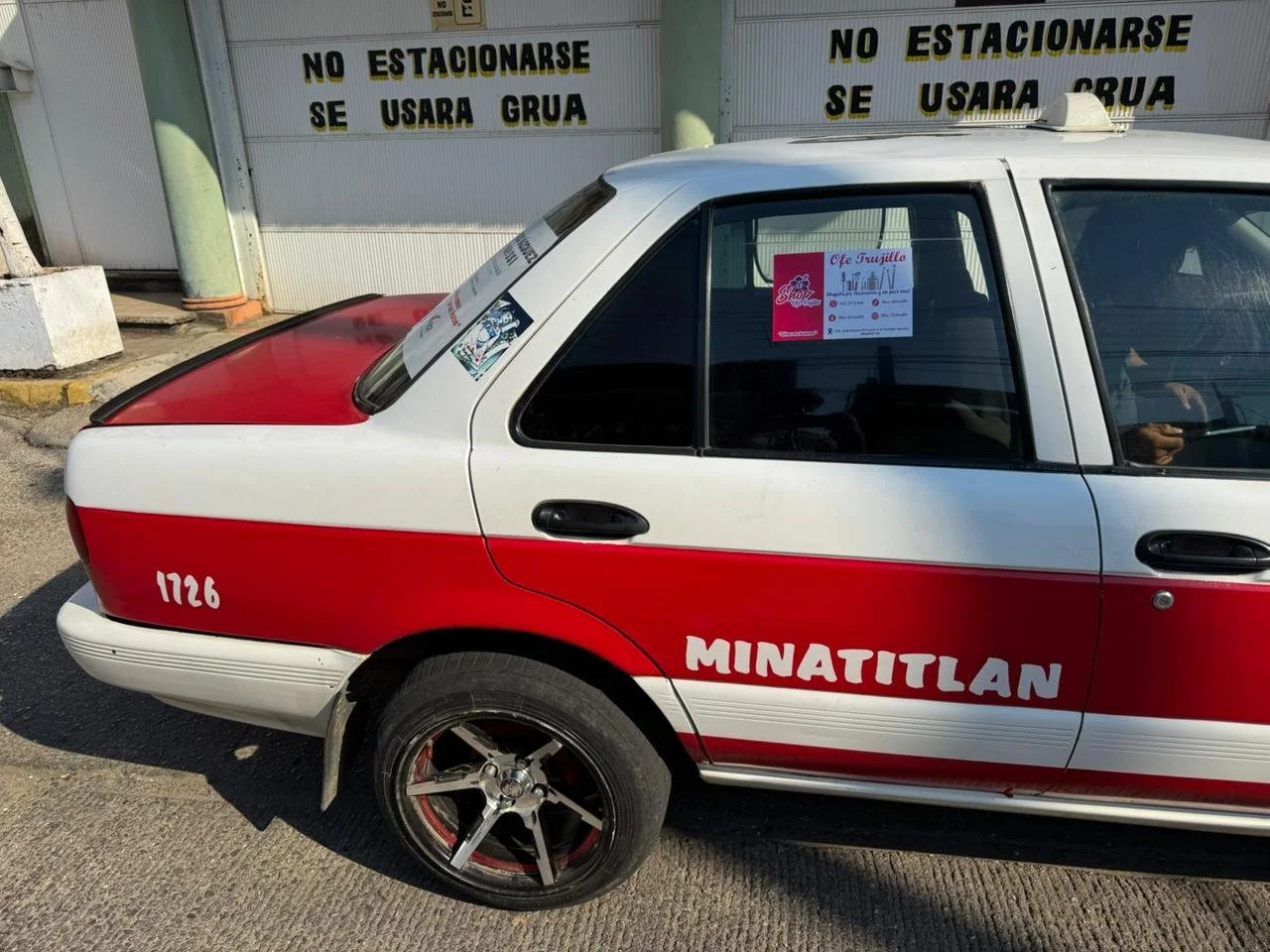 Auto de la policía de Minatitlán con matrícula 1726, color rojo y blanco, estacionado en la calle con señalamiento en fondo que dice 'No estacionarse, se usara grúa'.