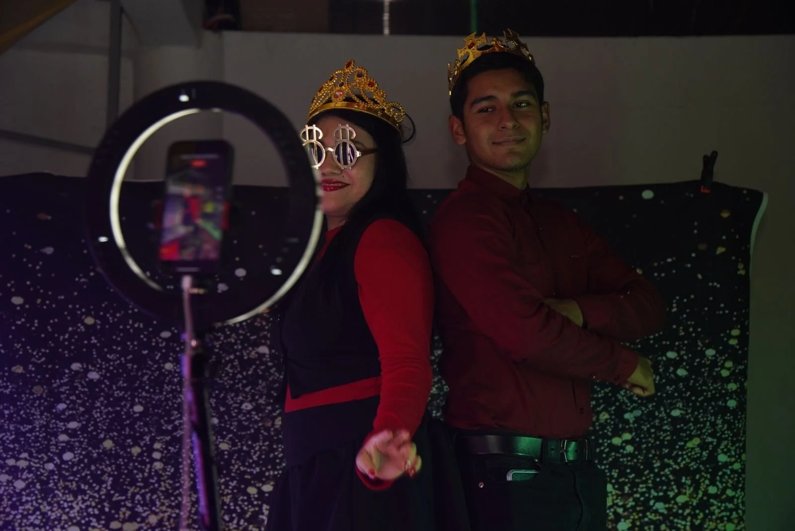 Dos personas con coronas sosteniéndose de las manos, una con gafas de dólares y un vestido negro y rojo y la otra con camisa blanca y pantalones oscuros, posando en un escenario con fondo de estrellas y partículas doradas.