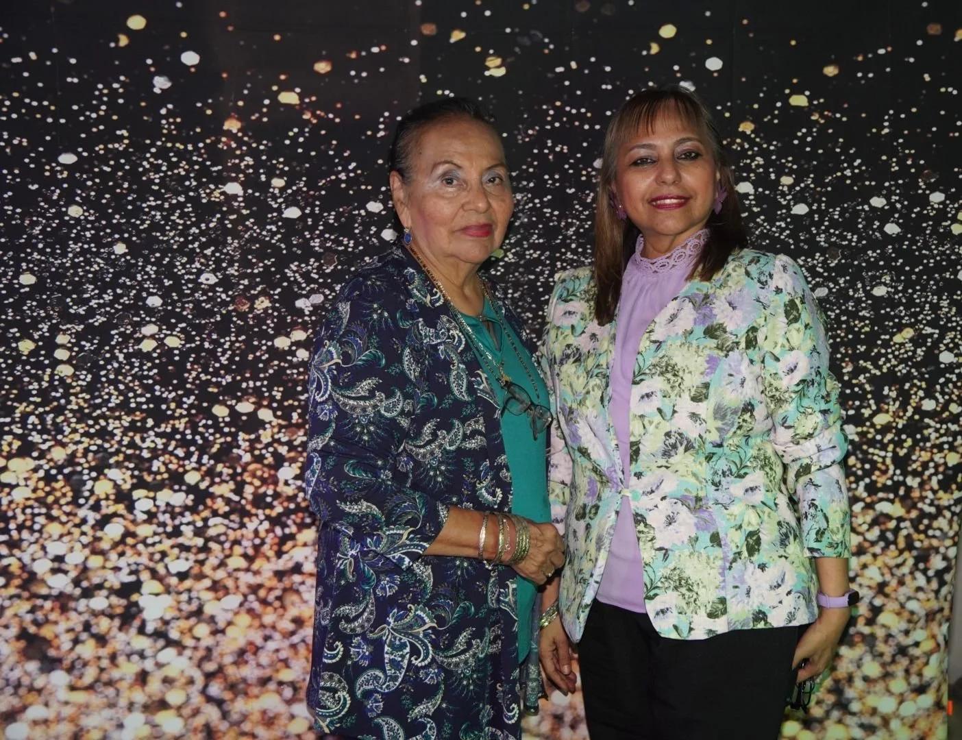 Dos mujeres posan juntas frente un fondo negro con puntos dorados que parecen luces o brillo, una de ellas lleva un vestido azul con patrones y accesorios dorados y la otra lleva una blusa lilac y una chaqueta de color claro con estampados.